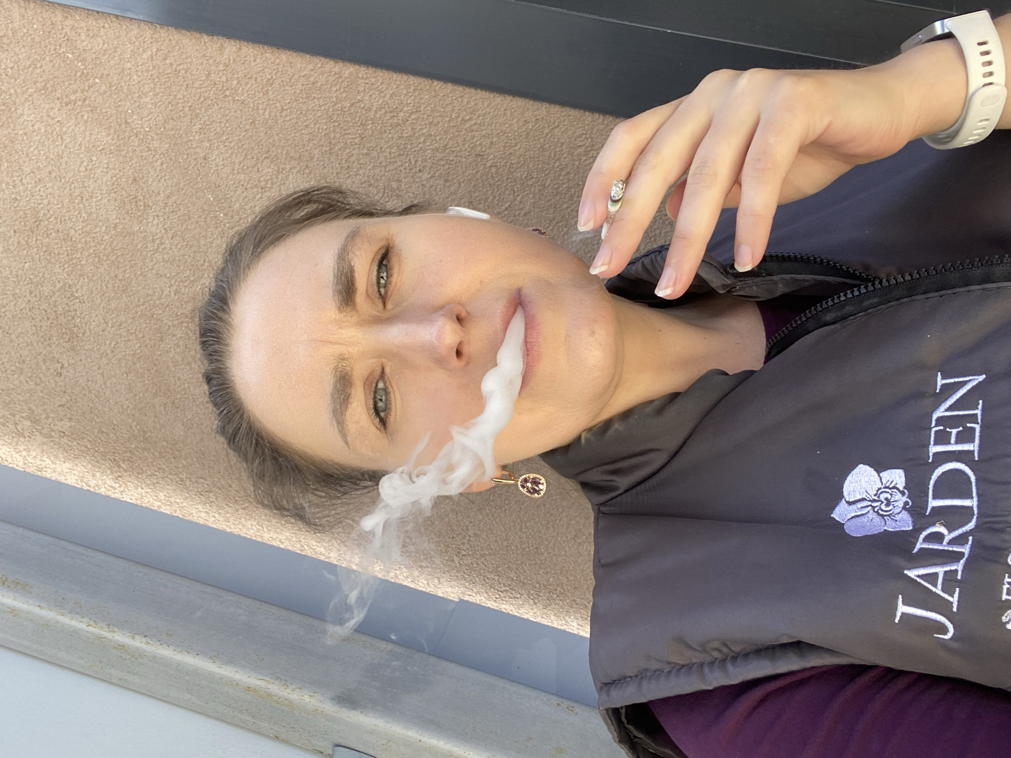 A woman is captured in a close-up, low-angle shot, exhaling a plume of white smoke from her mouth. Her head is tilted, and she gazes directly at the camera with blue-green eyes, her expression appearing focused or slightly squinting. Light brown hair frames her face. She holds a lit cigarette between the index and middle fingers of her right hand, which is adorned with a ring featuring a clear stone. On her left wrist, she wears a white smartwatch. A small, ornate earring is visible on her right ear, and a white earbud is in her left ear.
She is dressed in a dark grey or black puffer vest, beneath which a purple garment is visible. On the left side of her vest, the word "JARDEN" is embroidered in white capital letters, accompanied by a purple orchid logo.
The background consists primarily of a textured, light brown surface, possibly a wall or floor, with a darker horizontal element at the top. The lighting appears bright and natural, suggesting an outdoor or well-lit indoor setting. The scene takes place in Yekaterinburg, Russia.