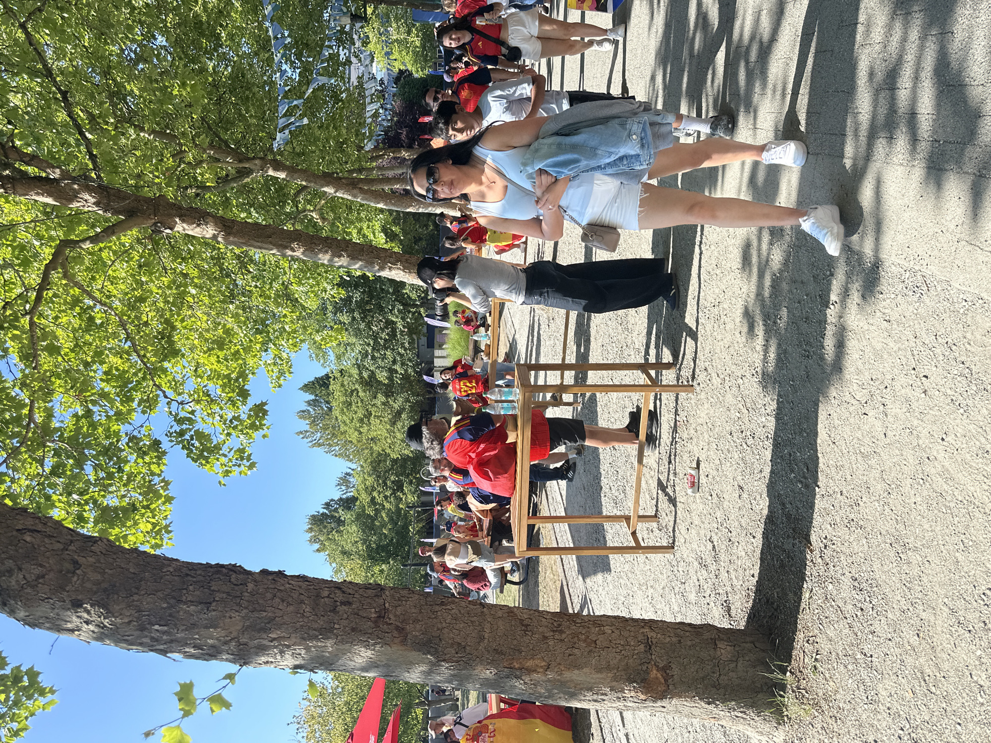 The image captures an outdoor scene on a sunny day, likely at a public gathering in Vancouver, Canada, given the context clues of Spanish flags and attire, possibly indicating a sporting event or cultural celebration. In the foreground, a woman in white shorts and a light blue tank top, holding a denim jacket, walks towards the viewer. Behind her, other people, many wearing red and yellow, are also walking and milling about. The environment is characterized by a gravelly ground, lined with trees that cast shadows. In the background, there are tables set up, some with people seated or standing around them, and decorative banners are strung between trees. One person is seen holding a camera, taking a picture. Red umbrellas are visible on the left side of the frame, suggesting a vendor or seating area. The overall atmosphere appears to be festive and casual.