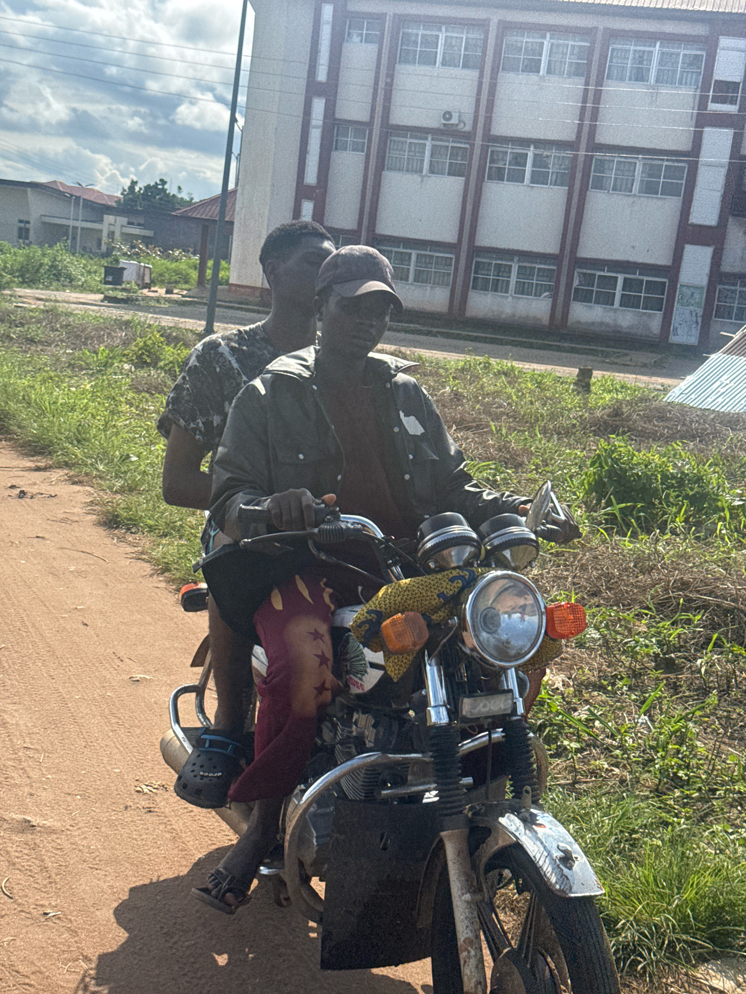 Two young Black males are riding on a motorcycle on a dirt road in Ihiagwa, Nigeria. The driver, wearing a dark jacket and a baseball cap, is operating the motorcycle. The passenger, wearing a camouflage t-shirt, is seated behind the driver. The motorcycle is decorated with colorful fabric and a red cloth hanging from the front. In the background, there are buildings, vegetation, and a cloudy sky. The scene appears to be set during the daytime.