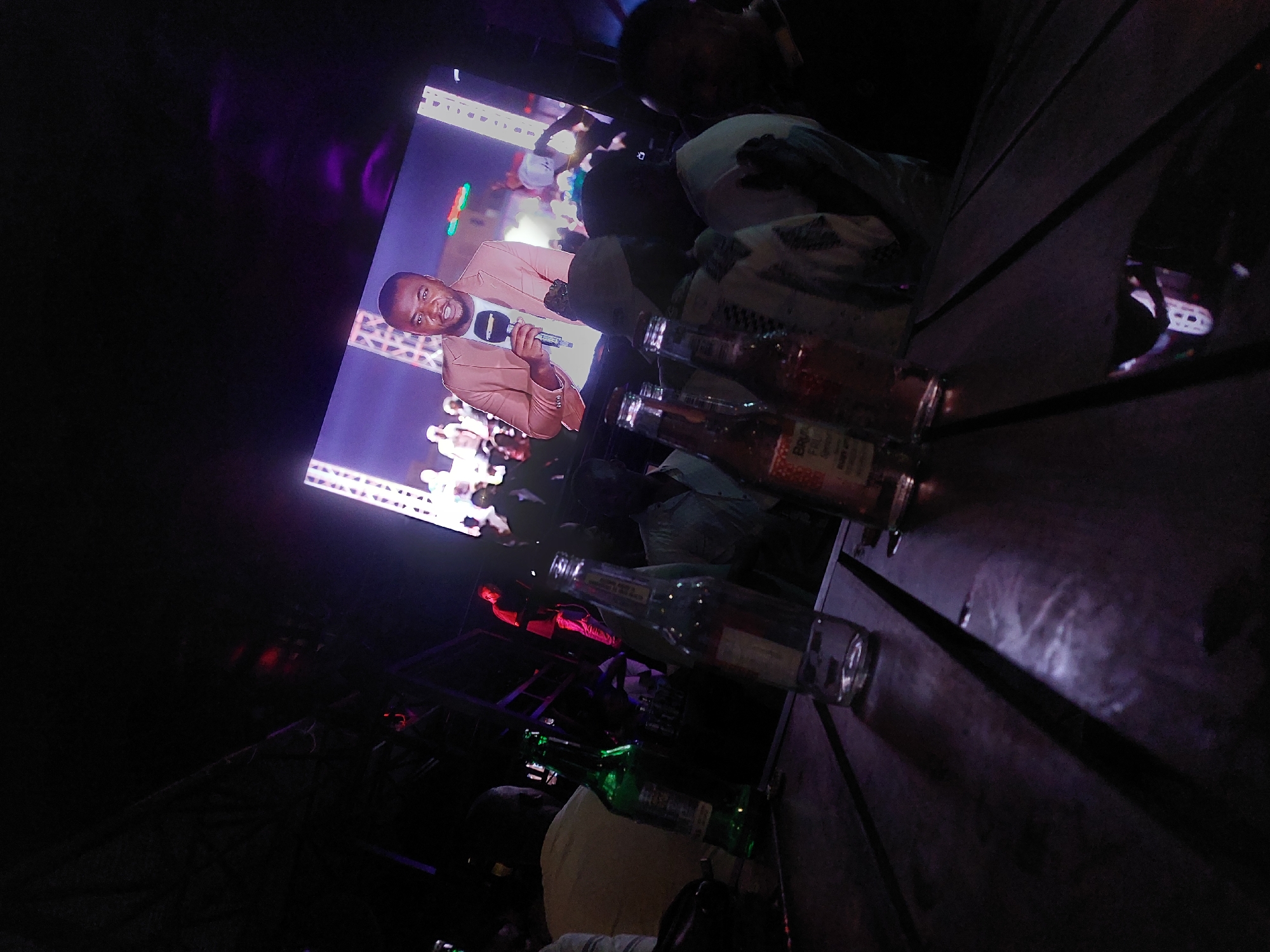 The image shows a dark indoor scene, likely a bar or club in Dar es-Salaam, Tanzania, based on the presence of local beer bottles on a wooden table. A television screen displays a man in a tan suit holding a microphone, possibly a live broadcast. Several people are present, though details are obscured by the dim lighting. Empty beer bottles are prominent on the table. The overall atmosphere suggests nightlife activity.
