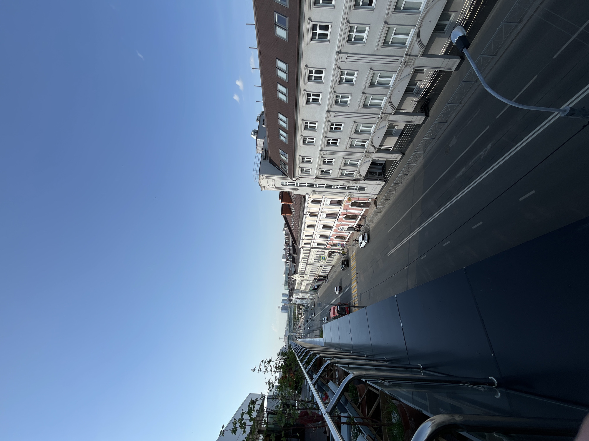 An elevated view looks down the street in Kazan, Russia. Buildings line both sides of the road. Cars drive on the street. A modern railing is in the foreground. The sky is blue with some clouds.