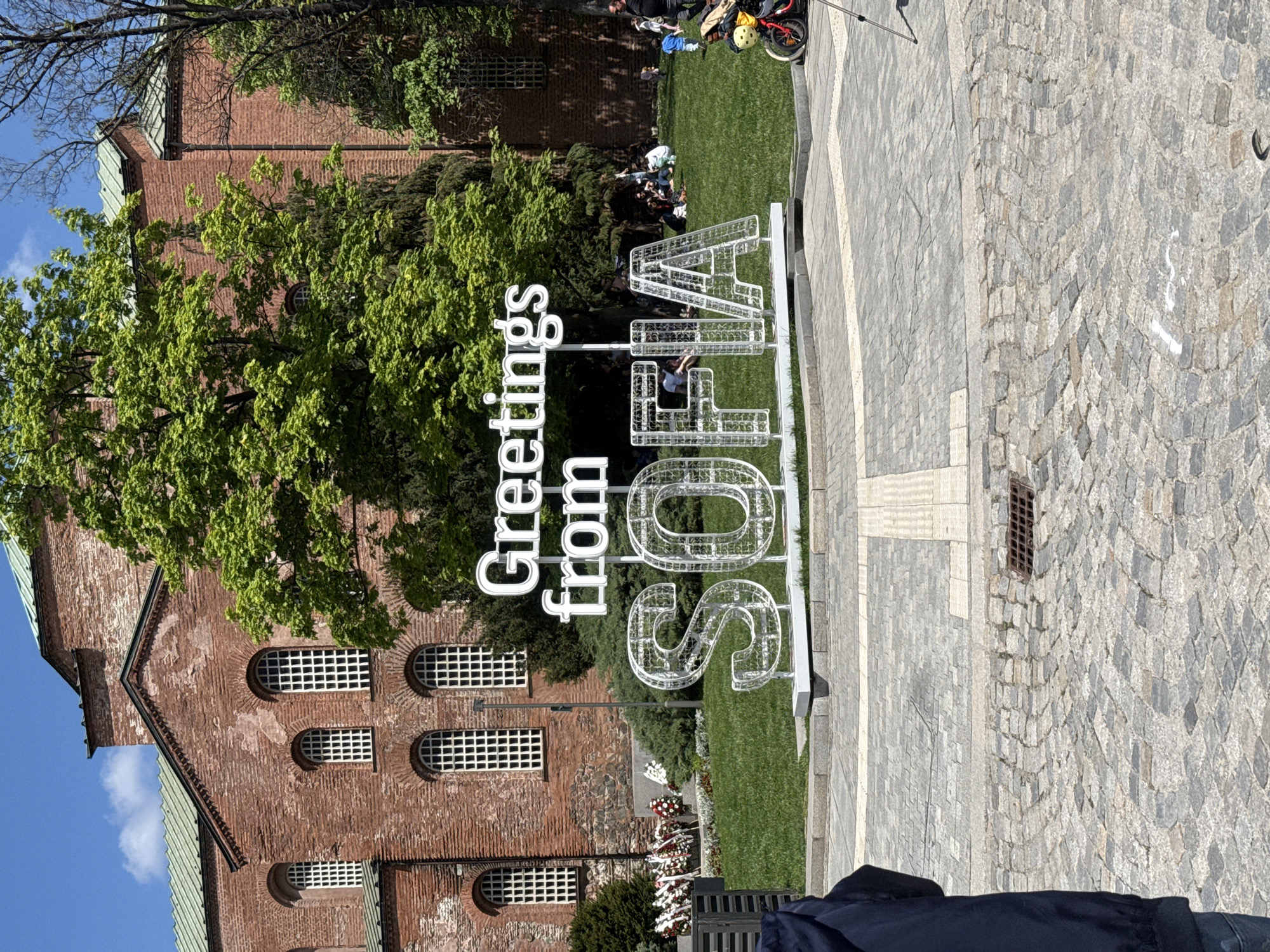 The image shows a public space in Sofia, Bulgaria. In the foreground is a sign that reads "Greetings from SOFIA" in large, white, wireframe letters. Behind the sign is a brick building with arched windows. A green tree partially obscures the view of the building. To the right of the sign is a patch of grass, and on the right is a paved area. People are visible in the background. The sky is blue with a few clouds. There are wreaths and floral tributes at the base of the sign.
