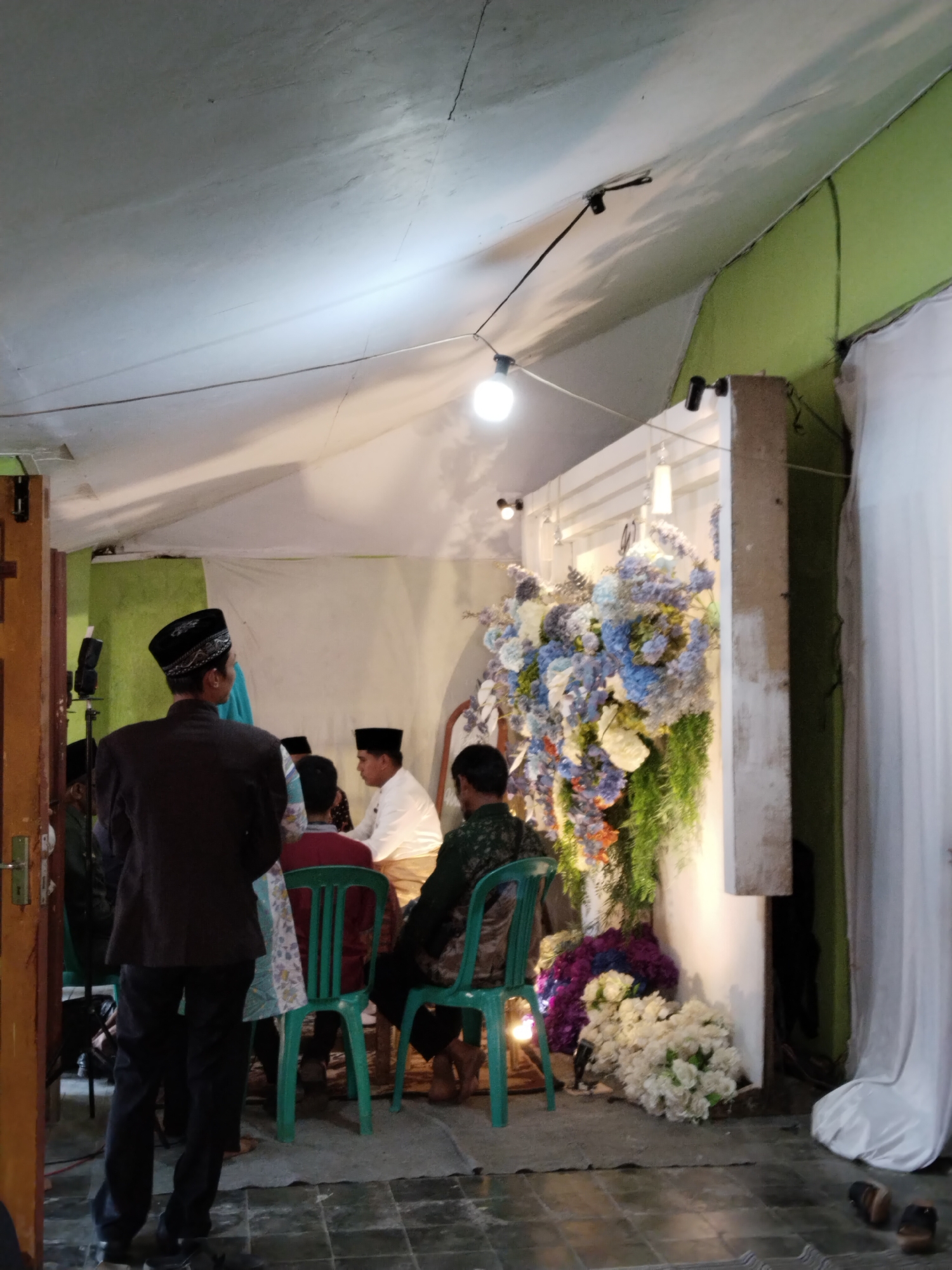 A group of people are gathered in a room, likely for a formal event, possibly a wedding ceremony or reception, in Kecamatan Pati, Indonesia. In the foreground, a man wearing a dark suit and a traditional black peci hat stands with his back to the camera, facing the group. To his right, a woman in a blue hijab is visible. The room's ceiling is white, with exposed wiring and lights. The walls appear to be painted a light color, with a section of bright green on the right.
The central focus of the scene is a decorative floral arrangement featuring an abundance of blue, white, and purple flowers, along with green foliage. The flowers are arranged against a white backdrop. Several individuals are seated on green plastic chairs, facing the floral display. One man in a white shirt and light-colored pants is seated prominently. Another man in a dark green patterned shirt is also seated. The floor is tiled with a dark grid pattern, and a gray rug is placed beneath the seated individuals. To the far right, white drapes hang, partially obscuring the wall. Shoes are visible on the floor in the lower right corner. The overall lighting in the room appears to be artificial, with several light sources contributing to the illumination.