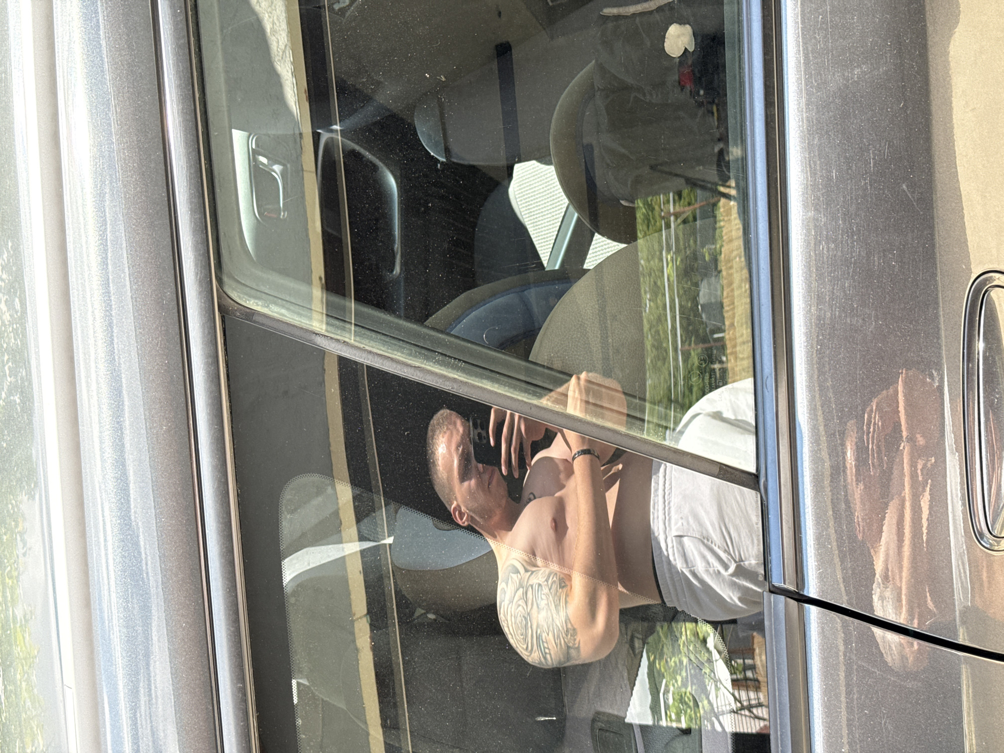 The image shows a man, shirtless and wearing white shorts, leaning out of a car window. He has extensive tattoos on his left arm and shoulder. He appears to be holding a smartphone in his right hand, possibly taking a selfie or a video. The interior of the car is visible, with seats and parts of the dashboard. The car's exterior is a metallic gray color. Outside the car, greenery and buildings are reflected in the window, suggesting an outdoor setting on a sunny day. The man's expression is neutral. The image is taken at an angle, with the man and car interior appearing rotated. There is some visible text on the car window, but it is illegible. The location is identified as Dobrogled, Bulgaria.