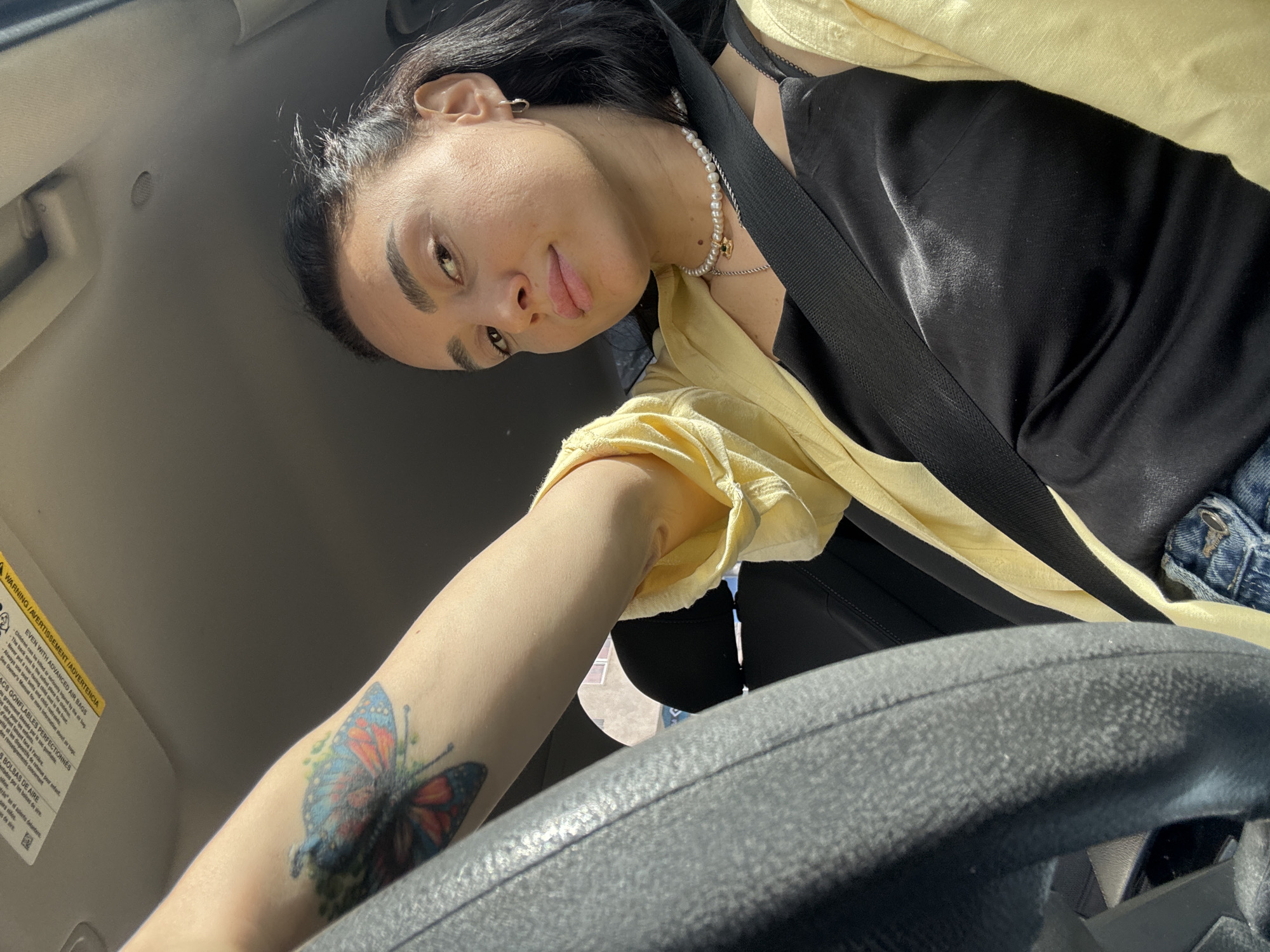 A selfie taken from inside a car. A woman is facing the camera and is seated with a seatbelt on. She wears a pearl necklace and a black satin top with a yellow jacket. The steering wheel is visible in the foreground, and her left arm with a butterfly tattoo is extended towards it. The car interior is visible, including the ceiling, door frame, and dashboard. Text indicating air bag warnings is visible on the upper left. The location is not discernible.