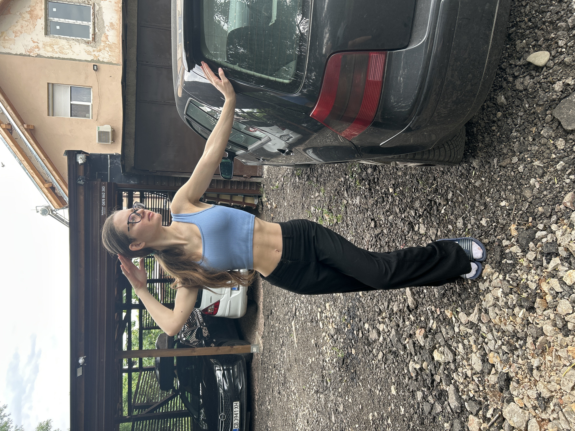 A single young Caucasian female, approximately 20-30 years old, stands in an unpaved yard composed of gravel and asphalt fragments, posing for the camera. She has long brown hair tied in a ponytail and wears dark-rimmed eyeglasses, a light blue ribbed crop top, black wide-leg trousers, white socks, and blue-and-white striped slides. Her midriff is exposed. Her left arm is raised with her hand near her head, while her right hand rests on the rear passenger side of a dark grey Volkswagen Golf Mk4 hatchback parked on her right.
In the background, to the left, a wooden carport shelters two vehicles: a black Opel Astra H displaying a Bulgarian license plate "CB 3941 XH" (indicating Sofia city registration) and, partially visible behind it, a white Opel Corsa. To the right of the carport, a two-story building with a light peach-colored, partially deteriorated facade is visible. This building features an external air conditioning unit, a window with a white roller shutter, and a solar water heater on its tiled roof. Below this building, a dark-colored structure, possibly a garage or workshop, displays a sign reading "NiKDoor 0878 862 383," consistent with a Bulgarian mobile number prefix. The environment is outdoors, under an overcast sky, within what appears to be a residential or private commercial property in Sofia, Bulgaria.
