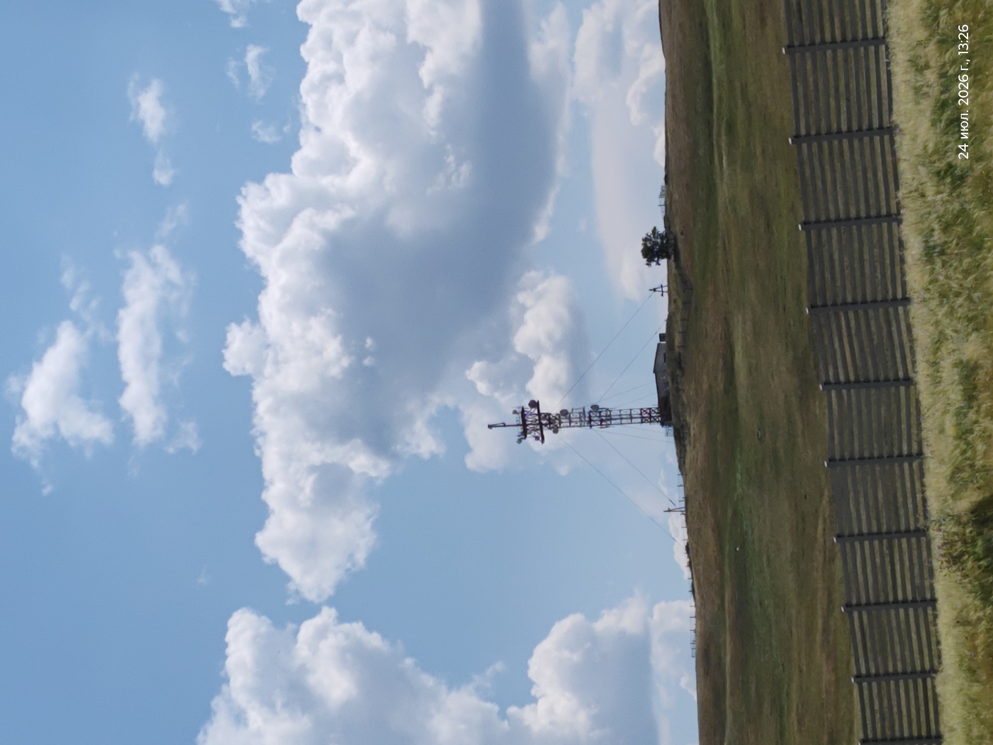 The image shows a telecommunications tower on a grassy hill under a partly cloudy sky. In the foreground, a wooden fence runs horizontally across the frame. The scene is located in Keneсары, Kazakhstan, as indicated by the date stamp "24 июл. 2026 г., 13:26" which translates to "July 24, 2026, 13:26". The tower is a lattice structure with multiple antennas and platforms. There are a few small buildings and a single tree visible on the hill near the tower. The grass on the hill is mostly green and brown, suggesting a summer or early autumn season. The sky is blue with scattered white cumulus clouds.