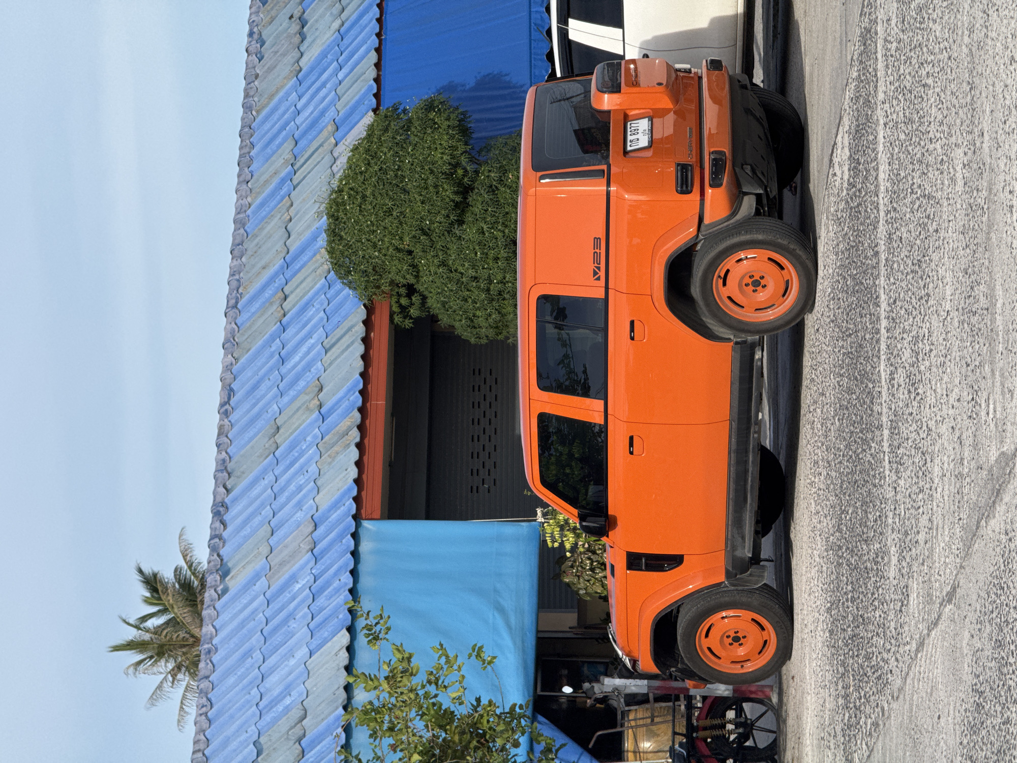 An orange, four-door SUV is parked on a concrete surface in front of a building with a blue, corrugated metal roof. The building also has a large blue awning extending from the front. To the left of the SUV, a portion of a red motorcycle is visible. The SUV has black trim around the windows and wheel wells, and its wheels are painted orange. A license plate on the rear of the SUV reads "กธ 8977" and "CHERY UGLIS". A small green tree with leaves and white flowers is growing on the right side of the building. A palm tree is visible behind the building's roofline on the left. The sky is clear and blue.