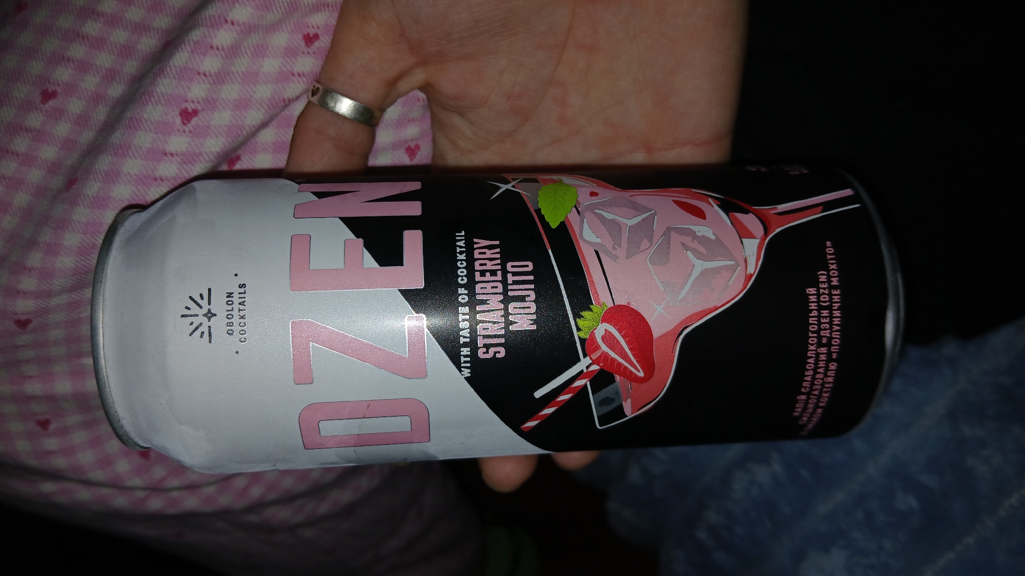 A hand holds a white and black can of "Dzen" Strawberry Mojito cocktail by Obolon Cocktails. The can features an illustration of a strawberry mojito drink. The background includes a pink and white patterned material and a silver ring on a finger. The can also has text in Ukrainian. The origin of this product is Ukraine. Dnipro is not known from this image.