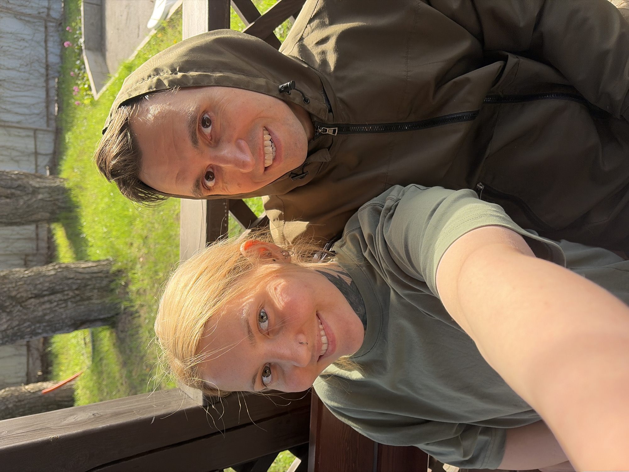 Two people, a man and a woman, are posing for a selfie outdoors. The man is wearing a dark green hooded jacket with the hood up. The woman is in a light green t-shirt. They are both smiling. The background shows trees, grass, and a wooden structure that appears to be a balcony or deck. The location is likely in Chaikovsky, Russia.