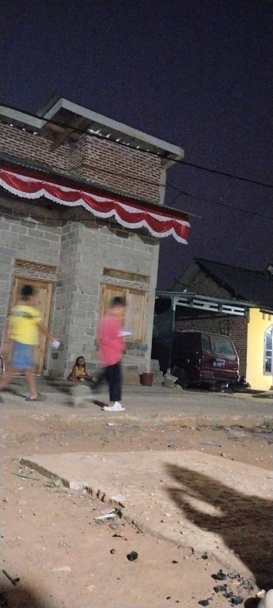 In Kali Balangan, Indonesia, at night, a brick and concrete building with a corrugated metal roof and a red and white draped awning stands on the left. In front of the building, two blurry figures are walking, one in a yellow shirt and blue shorts, the other in a red shirt and dark pants. A young child in a yellow dress sits on the steps. To the right, a maroon van is parked under an awning. The ground in front of the structures is unpaved, with visible debris and shadows cast by the figures. The sky is dark, indicating it is nighttime.