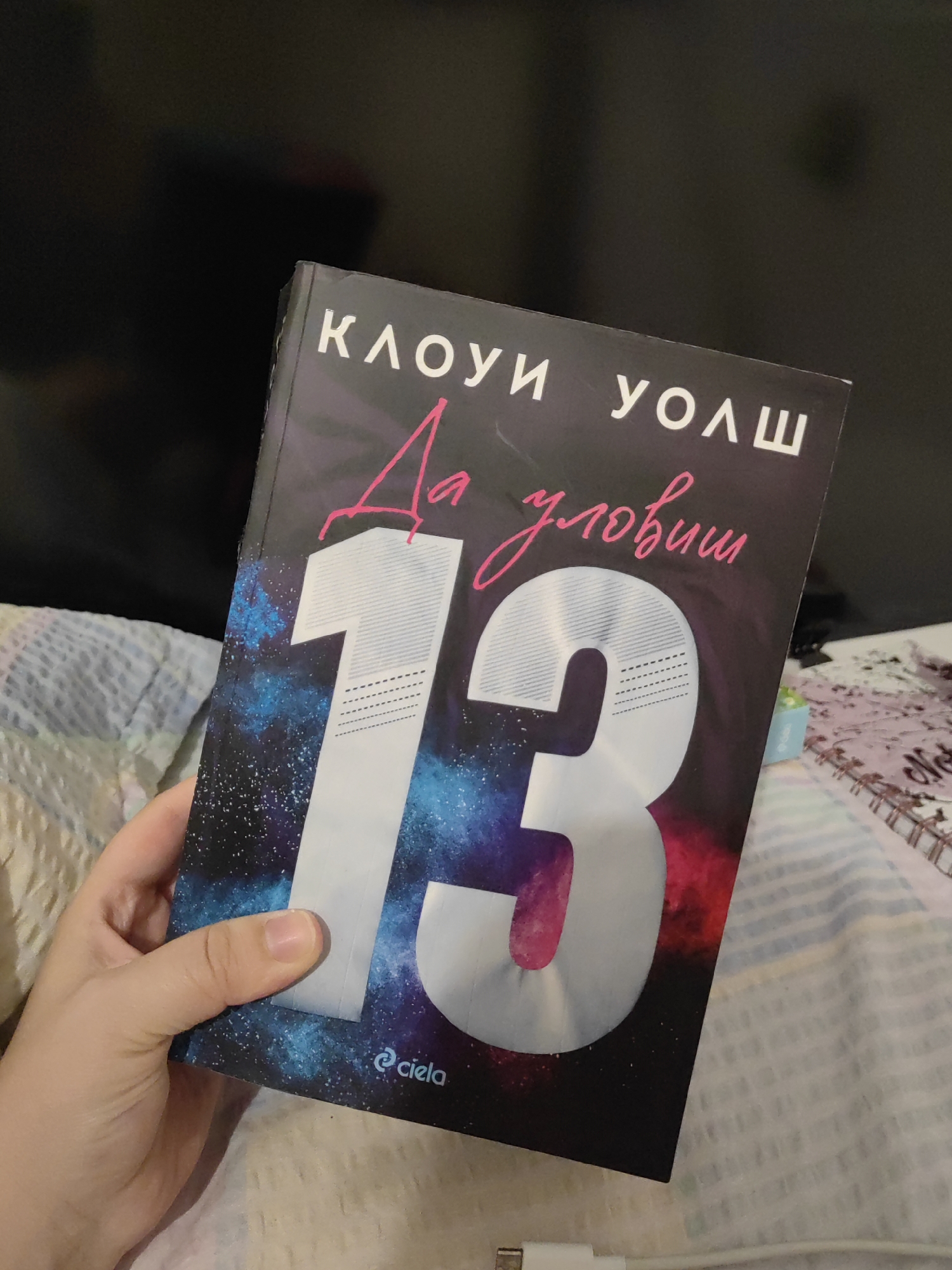A hand holds a book with a dark, galaxy-themed cover. White text at the top reads "КЛОУИ УОЛШ", and pink cursive text below that says "Да условишь". A large, stylized number "13" is prominently displayed in the center of the cover, rendered in silver with horizontal lines. At the bottom of the cover, a small logo with the text "© ciela" is visible. The book is held over a white and blue striped surface, possibly bedding. The background is dark and out of focus. This image does not contain any information related to the city of Djerovo, Bulgaria.