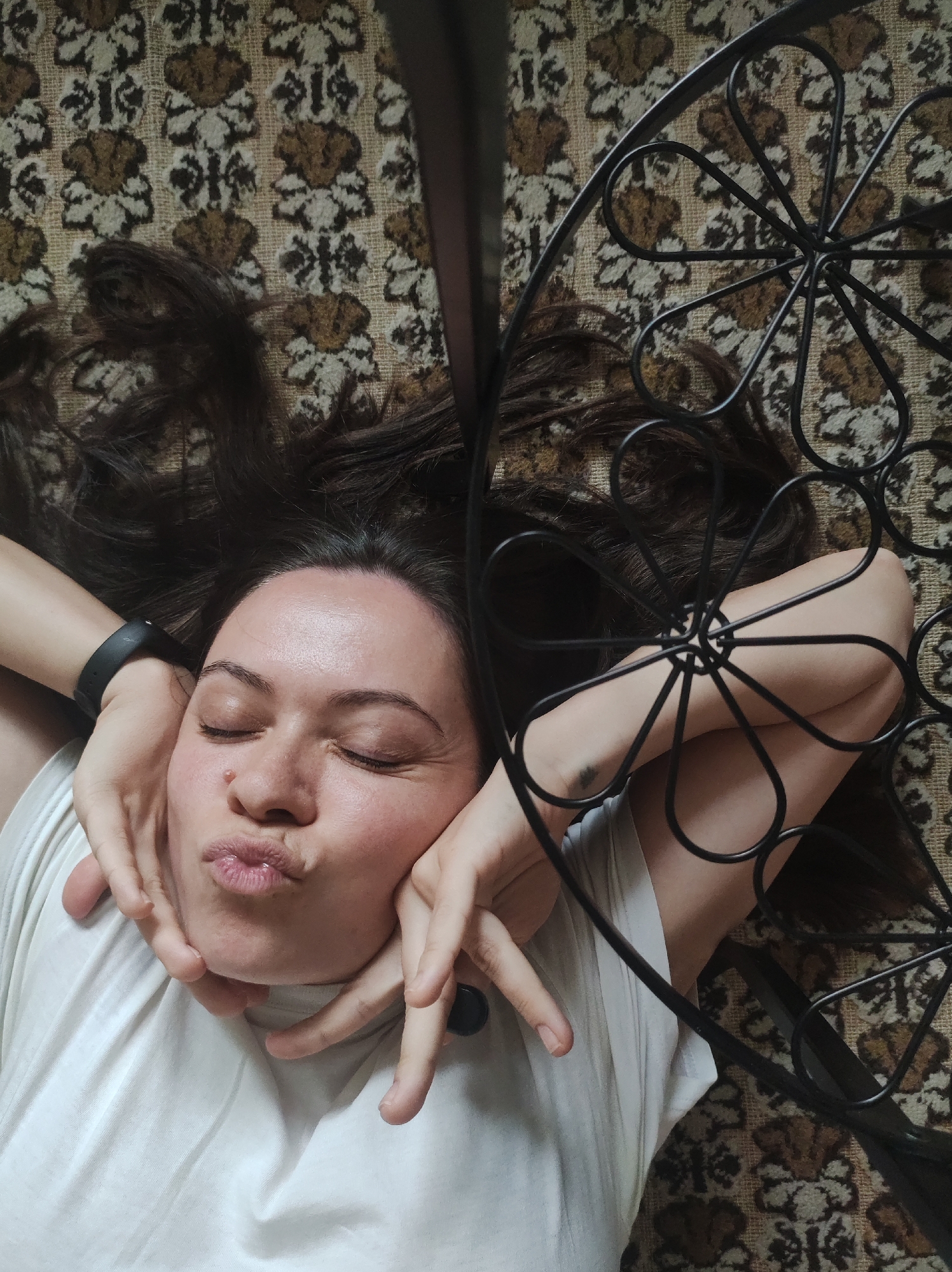 A person lies on a patterned carpet or rug, possibly indoors. The person's face is centered in the frame, eyes closed, with a pursed-lip expression. Dark hair spreads around the head, and hands are positioned on either side of the face. A black metal object with a floral design lies to the right of the person. The person wears a white shirt and a black wristband on their left wrist. The person has a beauty mark on the right side of their nose. There is a tattoo on the inner left forearm. The city of Фоки, Russia, cannot be determined from the image.