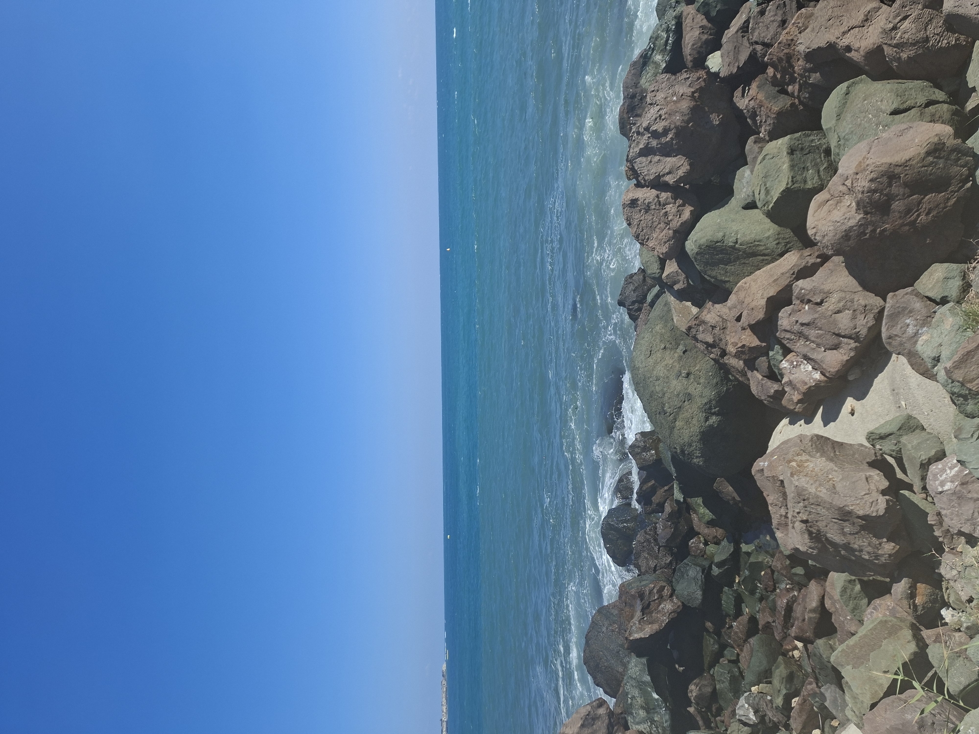 A wide-angle, eye-level, outdoor shot captures a bright, clear, blue sky above a choppy, turquoise ocean. The horizon line is visible, separating the sea from the sky. In the foreground, a jumble of large, weathered rocks of varying sizes and colors – ranging from deep brown and reddish hues to dark greens and grays – lines the shore. The waves crash against these rocks, creating white foam. In the distance, along the left side of the horizon, a faint line of buildings can be seen.