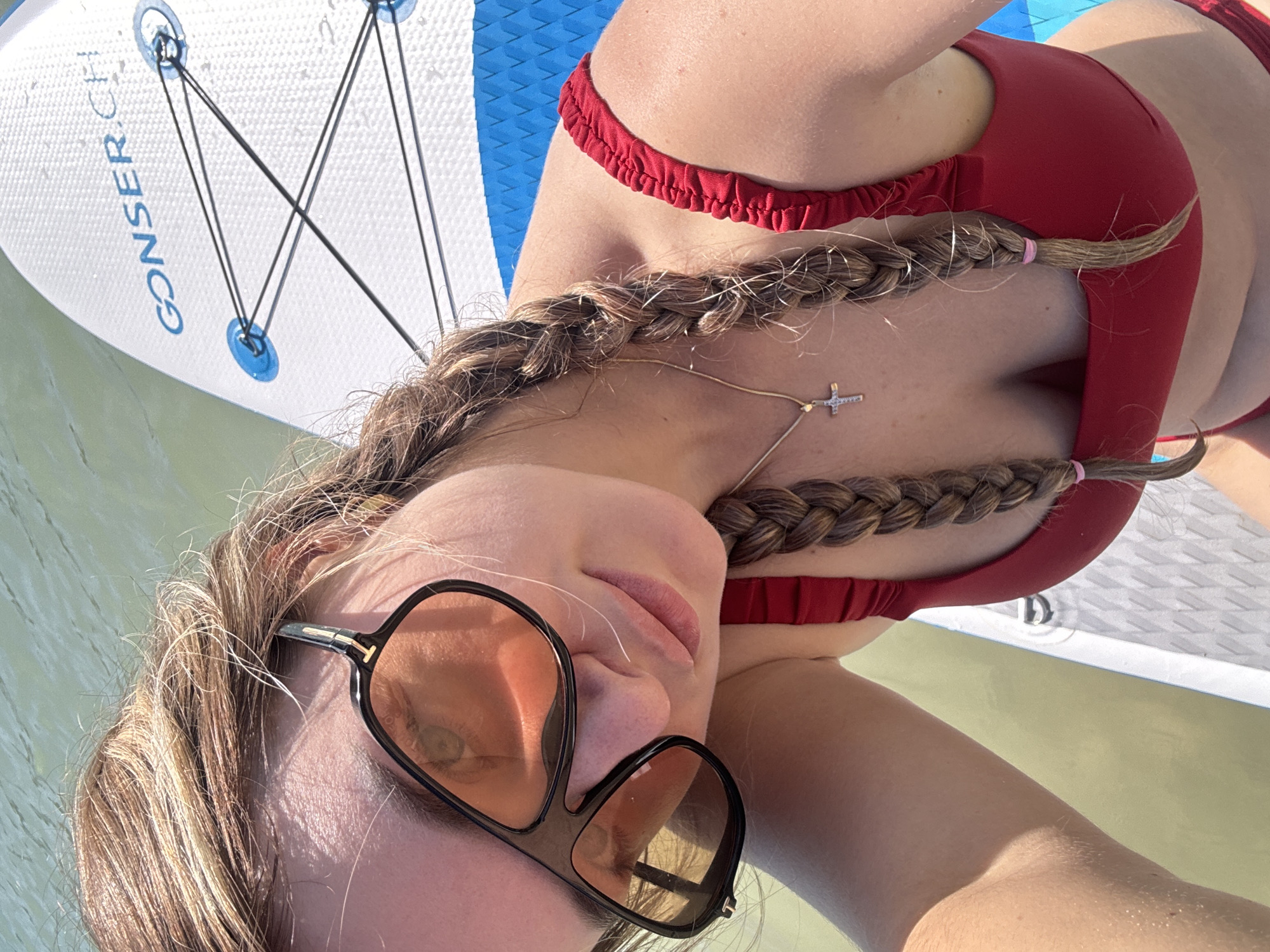 A young woman with blonde hair in two braids, wearing large sunglasses and a red bikini, is pictured in a close-up selfie. She is on a paddleboard in what appears to be a body of water. The paddleboard has the text "GONSER.CH" printed on it. The scene suggests an outdoor activity on a sunny day. The image does not provide information about the location in the city of Maisod, France.