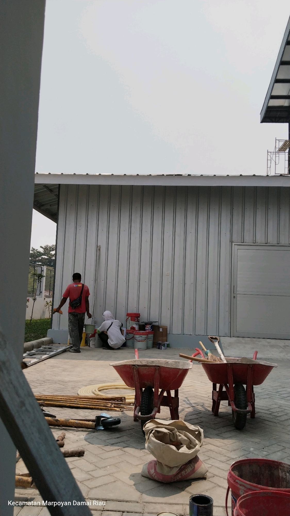 A construction site in Marpoyan Damai District, Pekanbaru, Riau Province, Indonesia. Two workers are present, one in a red shirt and black fanny pack, holding a trowel, and another in white, kneeling and working with paint cans. Several large red paint buckets, smaller paint containers, and some cardboard boxes are arranged near the kneeling worker. Two red wheelbarrows are positioned in the foreground, one empty and the other containing dirt and a broom. Various construction materials, including pipes, rebar, and a grinding tool, are scattered on the paved ground. A light-colored corrugated metal building with a gray door stands in the background, with a portion of a metal roof structure visible above. The sky is overcast.