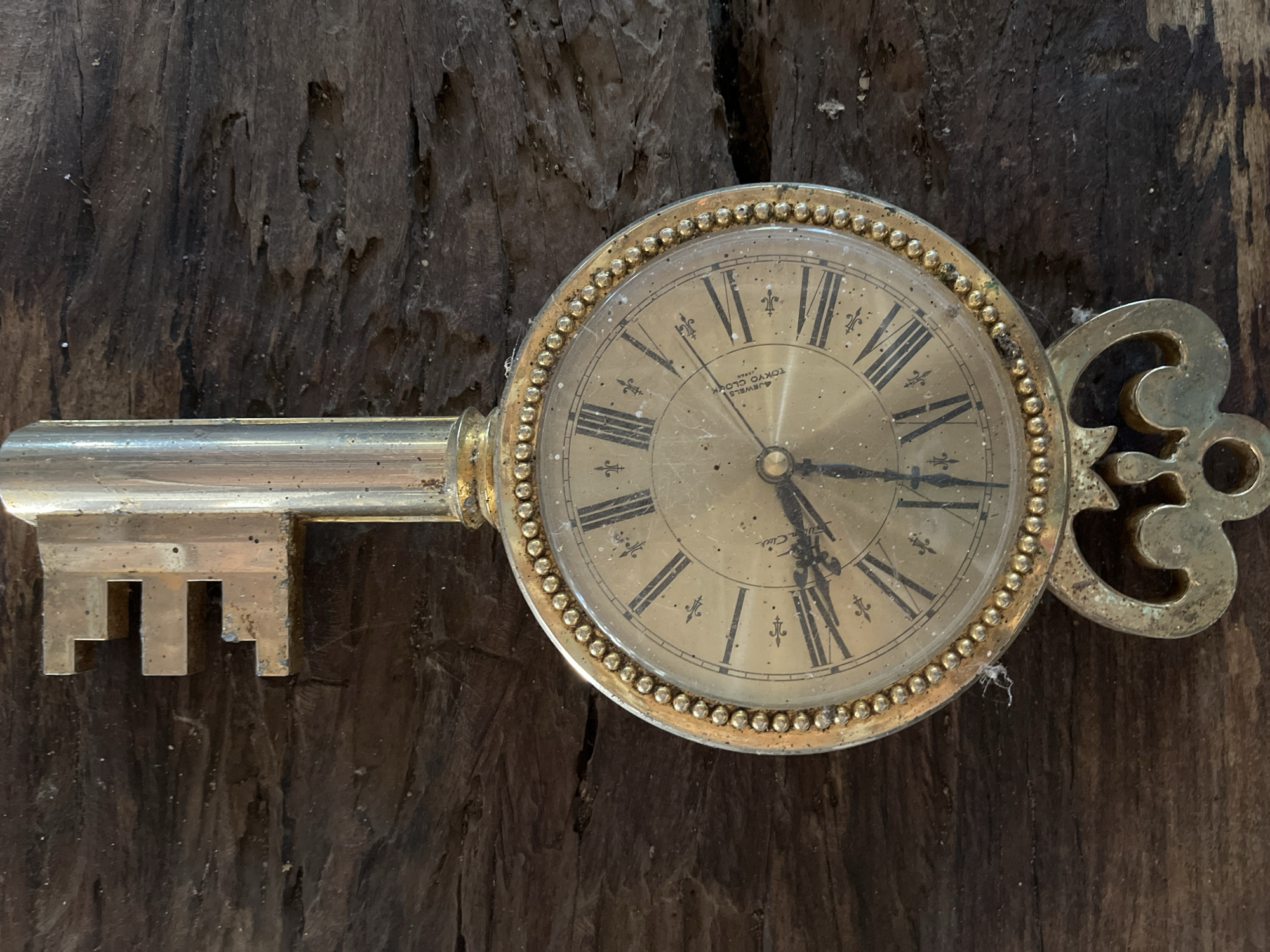 A decorative, antique brass key-shaped clock is centrally positioned against a dark, weathered wooden background. The clock face features Roman numerals and ornate hands, with the time appearing to be approximately 3:53. The key's head is at the top, and the ornate key bit is at the bottom. The wooden surface exhibits a rough, textured grain and appears to be part of an outdoor structure, possibly a wall or table, in the city of Tambakreja, Indonesia. There are no people or animals visible in the image, and no discernable actions are taking place beyond the stationary display of the clock and the weathered environment.