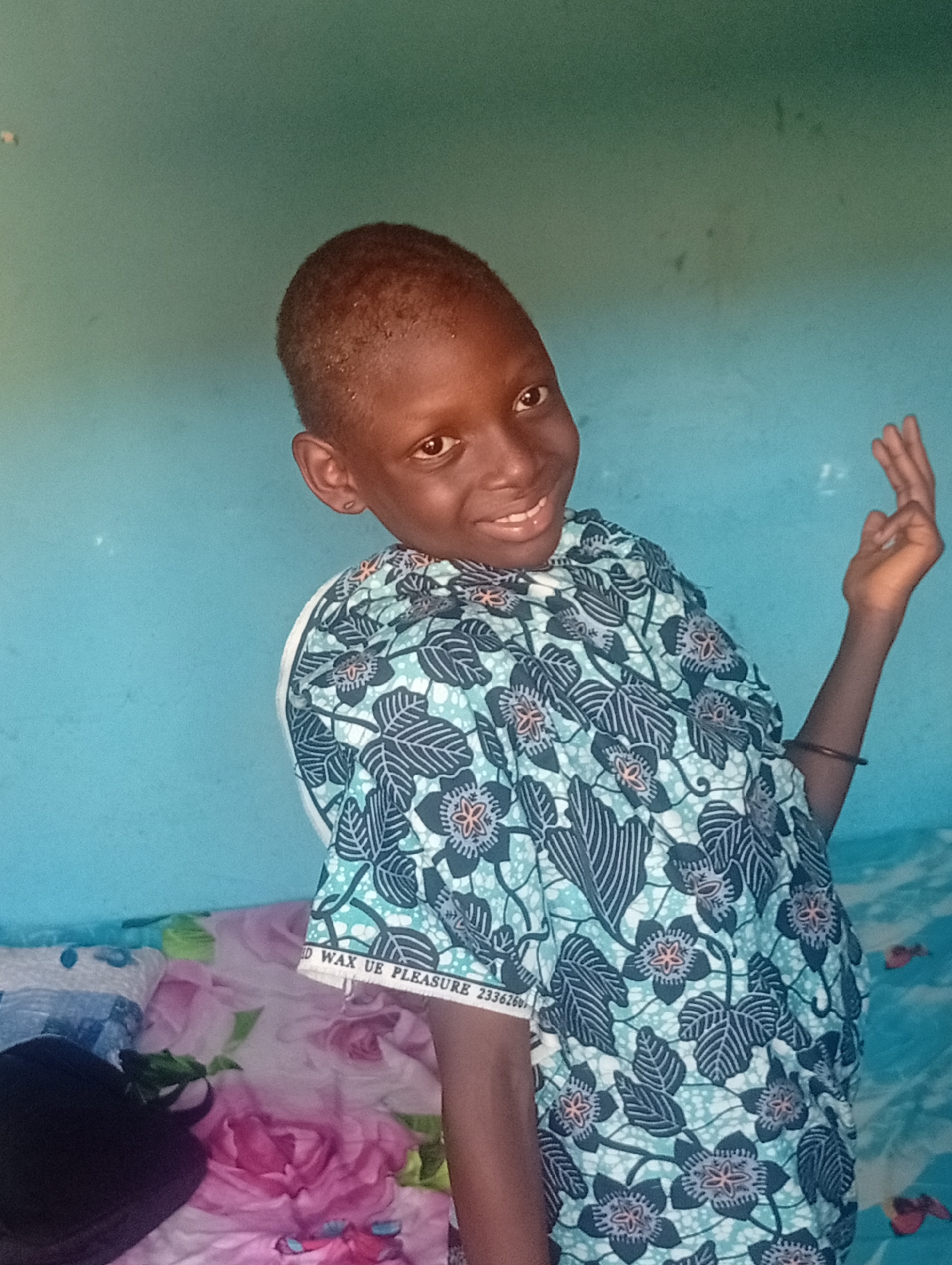 A young child stands indoors against a light blue wall, dressed in a blue patterned shirt. They are smiling and holding their right hand up, fingers slightly spread. The background includes a bed with floral-patterned bedding. The location is Unknown, Unknown.