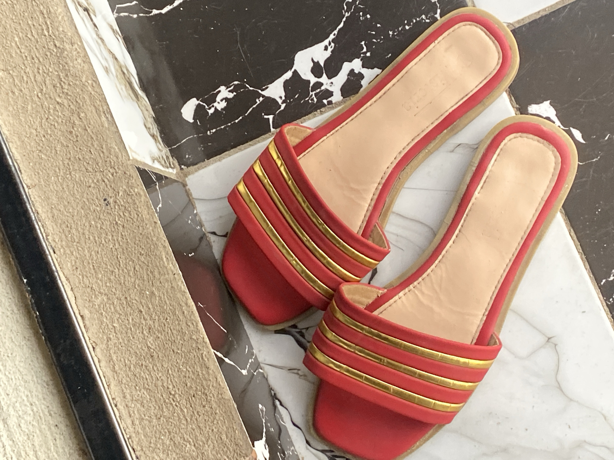 The image shows a pair of red slide sandals with gold trim, resting on a tiled floor. The floor has a black and white marble pattern. The sandals are positioned diagonally across the frame, with the left sandal slightly closer to the viewer. The background consists of a textured, light-colored surface with a dark metallic strip running along the top edge. The overall scene suggests an indoor setting, possibly a home or a shop, located in Jauro Yinu, Nigeria.