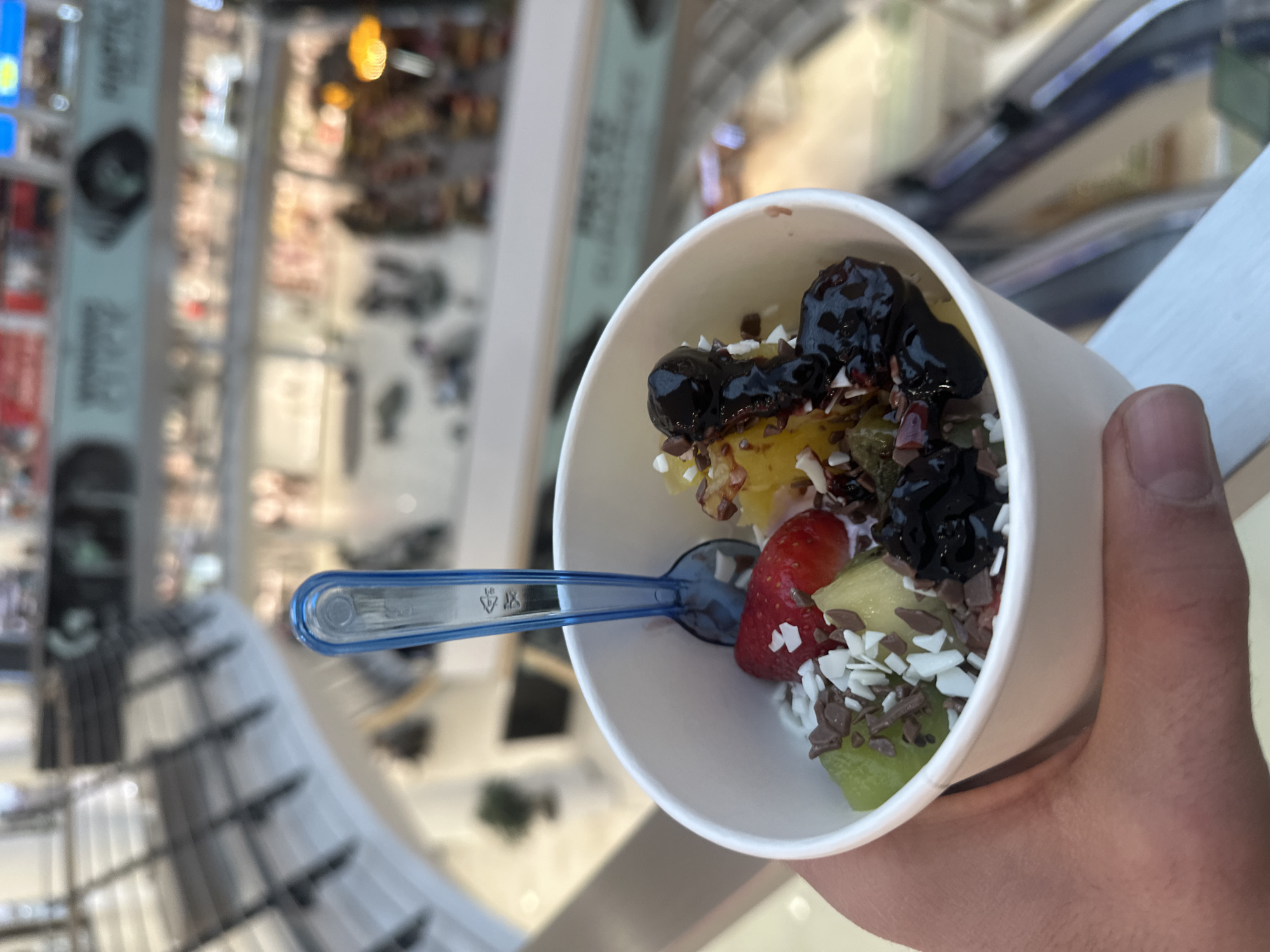 An individual's light-skinned hand holds a white paper cup containing a dessert, presumably frozen yogurt, topped with various ingredients. Visible toppings include diced green kiwi, diced yellow fruit (likely pineapple or mango), a single red strawberry, dark berries (such as blueberries or blackcurrants) coated in syrup, and both dark and white chocolate shavings or sprinkles. A translucent blue plastic spoon is embedded upright in the dessert. The scene is set indoors, within a multi-level shopping mall in Sofia, Bulgaria. The background, blurred due to a shallow depth of field, reveals curved white architectural railings, blurred escalators or stairs, and indistinct retail storefronts with illuminated signage on multiple floors. Several blurred figures of people are discernible on the lower levels, indicating a populated commercial environment.