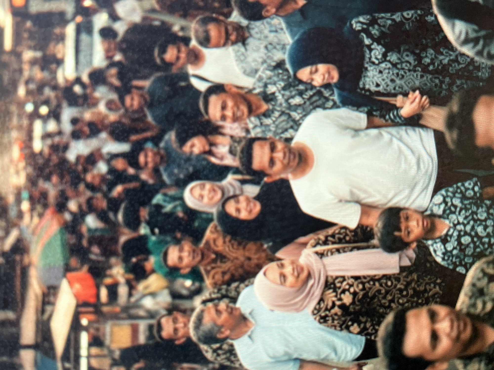 The image captures a crowded street scene in Sejahtera, Indonesia, likely a bustling market or thoroughfare. A dense throng of people, predominantly adults with a few children, are visible, navigating the crowded space. Many individuals are wearing traditional Indonesian attire, including batik shirts and headscarves, indicating cultural significance. The environment appears to be urban, with stalls and makeshift structures lining the street, suggesting a commercial area. The lighting indicates it is daytime, with some areas of shadow and highlight. The overall atmosphere is one of lively activity and pedestrian movement.