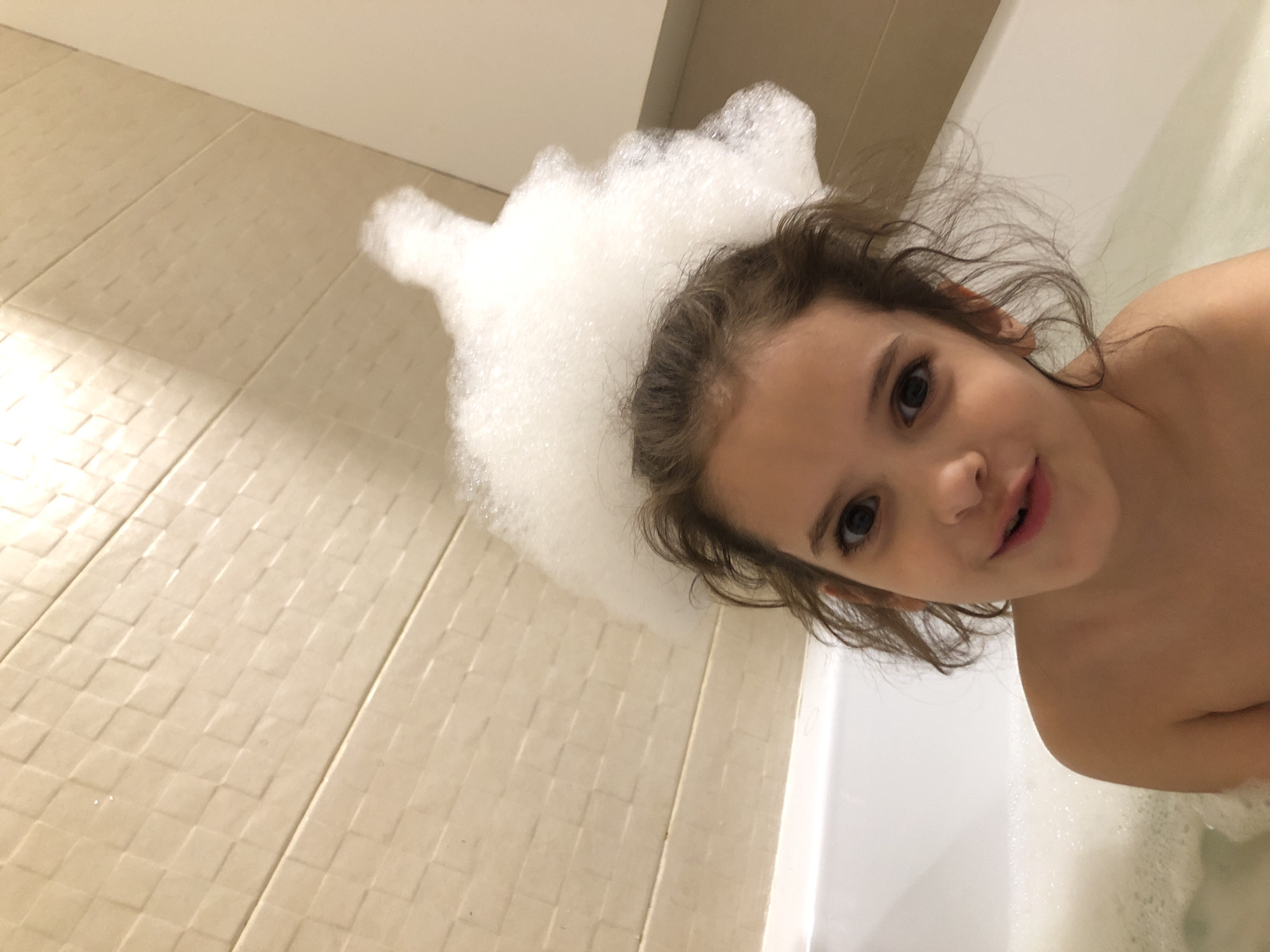 An eye-level shot captures a young child, likely a girl, taking a bubble bath in a modern bathroom. The child is positioned in the bathtub, looking directly at the camera with an engaging gaze and a soft, gentle smile. Her dark, wavy hair is damp and frames her face, while a large, frothy mass of white bath bubbles covers a significant portion of her head and shoulders, extending over the edge of the tub onto the tiled floor.
The bathroom floor is covered with light-colored, square tiles that feature a subtle, textured pattern, possibly a small mosaic or basketweave design. The tiles are a neutral beige or cream color, and warm, artificial lighting casts soft shadows across them. The bathtub is white and clean, with more bubbles visible in the water around the child.
The overall impression is one of a cheerful and innocent moment during a bath. The photograph's angle is slightly tilted, with the child's face and upper body dominating the right side of the frame and the tiled floor occupying the left. There is no discernible text visible in the image. The location context for this scene is Saint Petersburg, Russia.