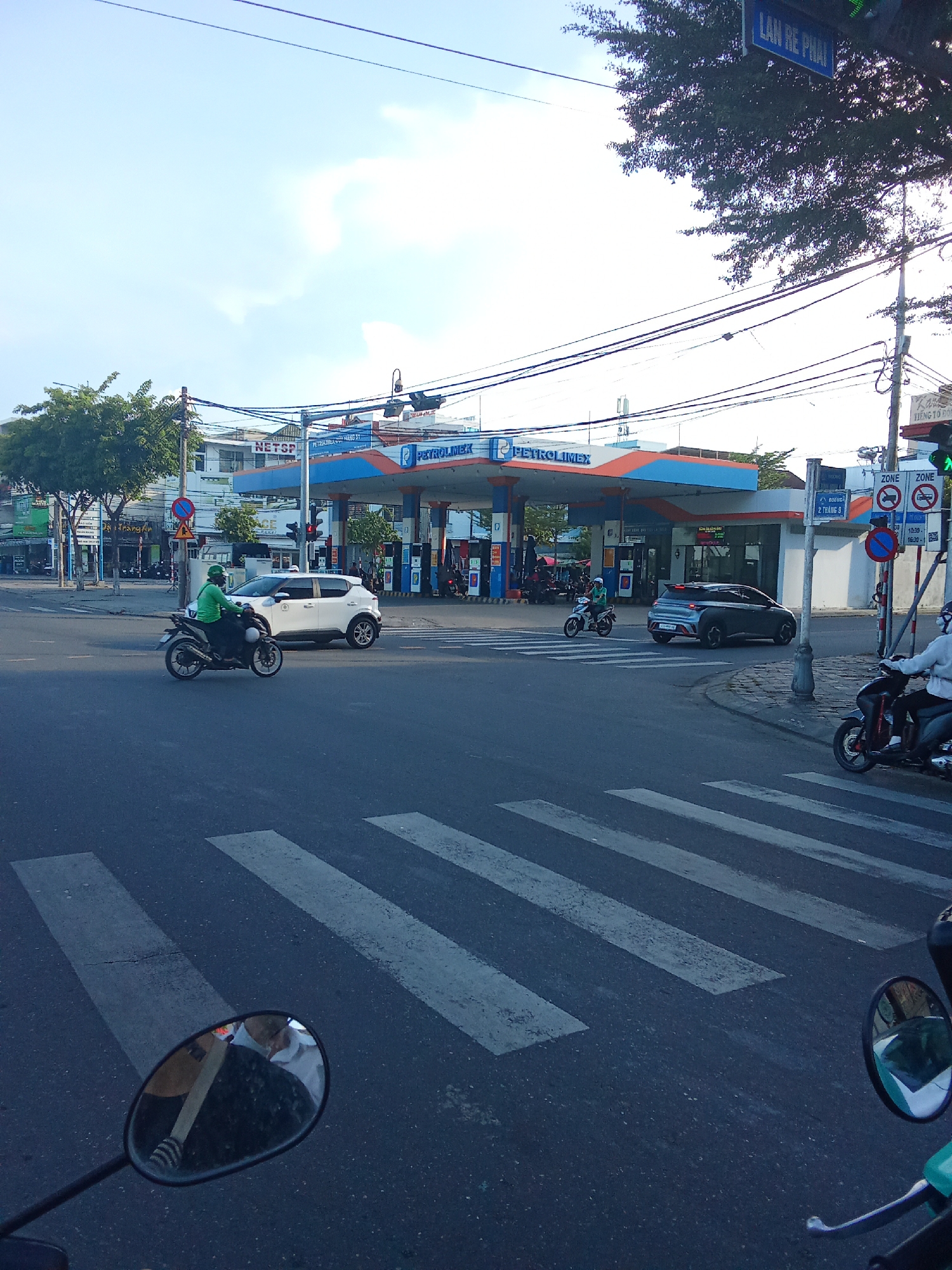 The image depicts a street intersection in Da Nang, Vietnam, featuring a Petrolimex gas station. A white car and a motorcycle are stopped at a crosswalk, with another motorcycle passing through. The gas station has prominent "PETROLIMEX" branding. Overhead power lines are visible, along with street signs, including one that reads "LAN RE PHAI." The sky is partly cloudy. The perspective suggests the photo was taken from another motorcycle, with a rearview mirror reflecting the rider and the scene.