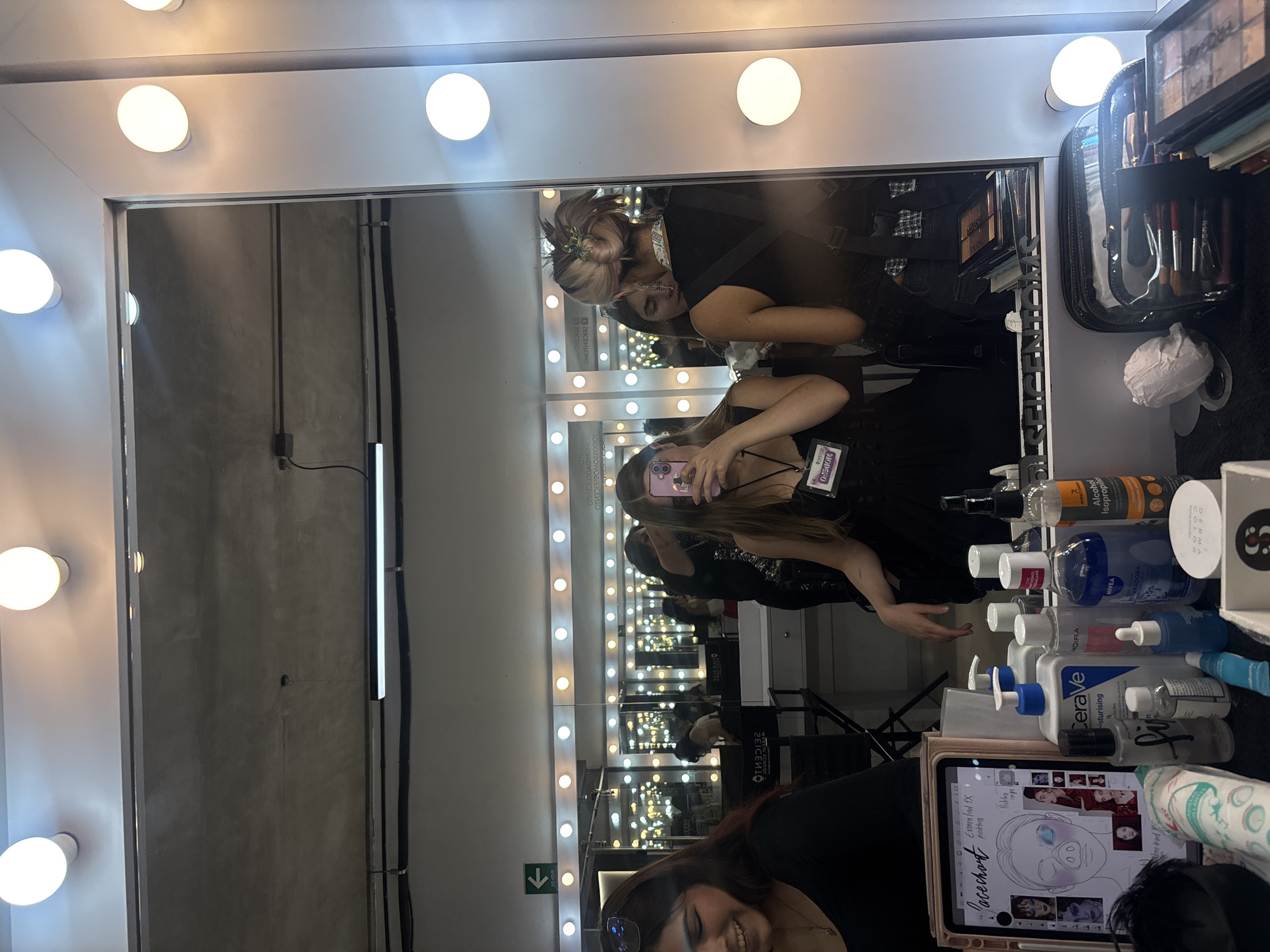 The image, taken in Mexico City, Mexico, features a reflection in a vanity mirror with surrounding lights. Two women are visible in the mirror. One woman is holding a phone, presumably taking the photo. A table in front of the mirror holds an array of makeup products, including CeraVe moisturising lotion, Moira face wipes, Nivea lotion, alcohol and derma color brand. A tablet is visible, displaying an image relating to makeup and design. In the background, the wall is gray and has electrical wiring visible. The room is lit by the vanity lights.