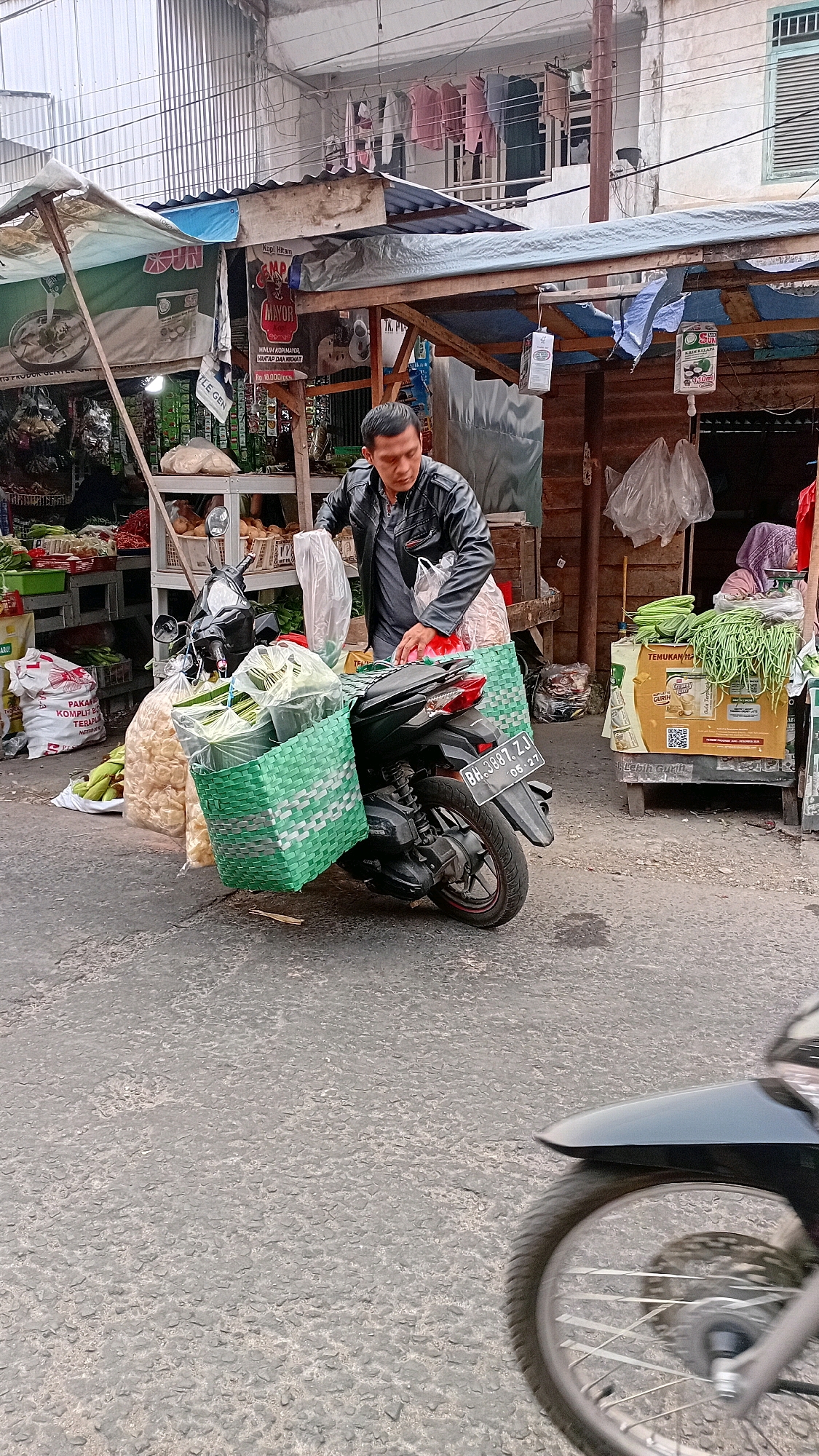 A man in a black leather jacket is standing next to a motorcycle loaded with green woven baskets filled with produce. The motorcycle's license plate reads "BH 3887 ZJ". The scene appears to be a market or roadside stall in Jambi, Indonesia, with various goods and structures visible in the background. A woman in a headscarf can be seen in the background at a stall. Clothes are hanging on a line overhead.