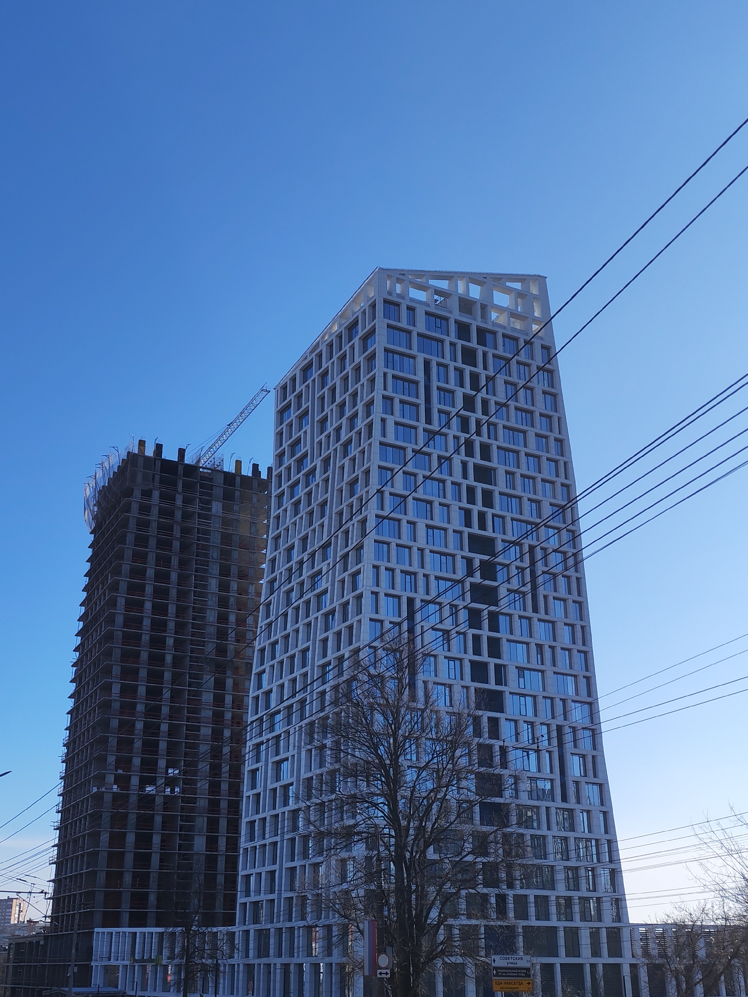 The image shows two tall buildings in Izhevsk, Russia under a clear blue sky. The building on the right is a modern high-rise with a repetitive grid pattern of windows. It appears to be completed, with a decorative top. A bare tree stands in front of it. To the left is a building under construction, with exposed concrete framework and a crane at the top. Power lines run diagonally across the upper right portion of the image. A low barrier or wall is visible in the foreground. The area appears to be an urban environment. The sign indicating "SOVETSKAYA" provides the location in the city of Izhevsk, Russia.