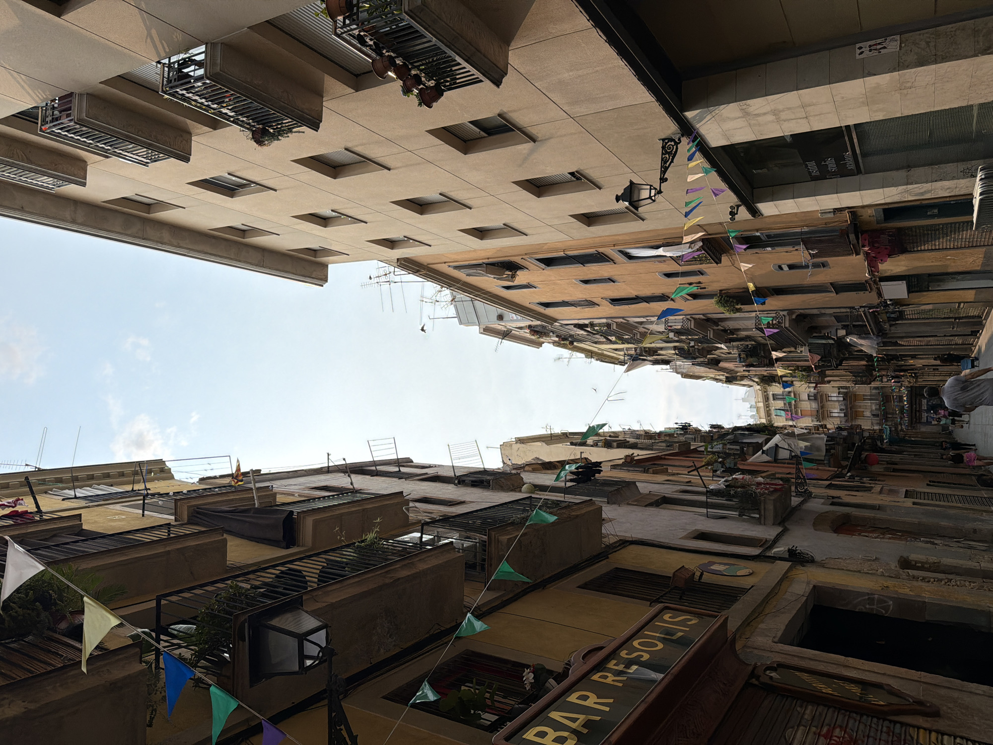 This is a low-angle, wide shot of a narrow street in Barcelona, Spain. The street is lined with tall, multi-story buildings on both sides, casting shadows and creating a sense of enclosure. Colorful pennant flags are strung across the street, adding a festive atmosphere. On the left side of the street, a building with a yellow facade features balconies with black railings and planters. A sign reads "BAR RESOLIS". On the right side, a building with a beige facade also has balconies with railings and planters. The sky above is a pale blue with scattered clouds. The street is paved and appears to be a pedestrian walkway, with a few people visible walking in the distance. The architecture suggests an older European city.