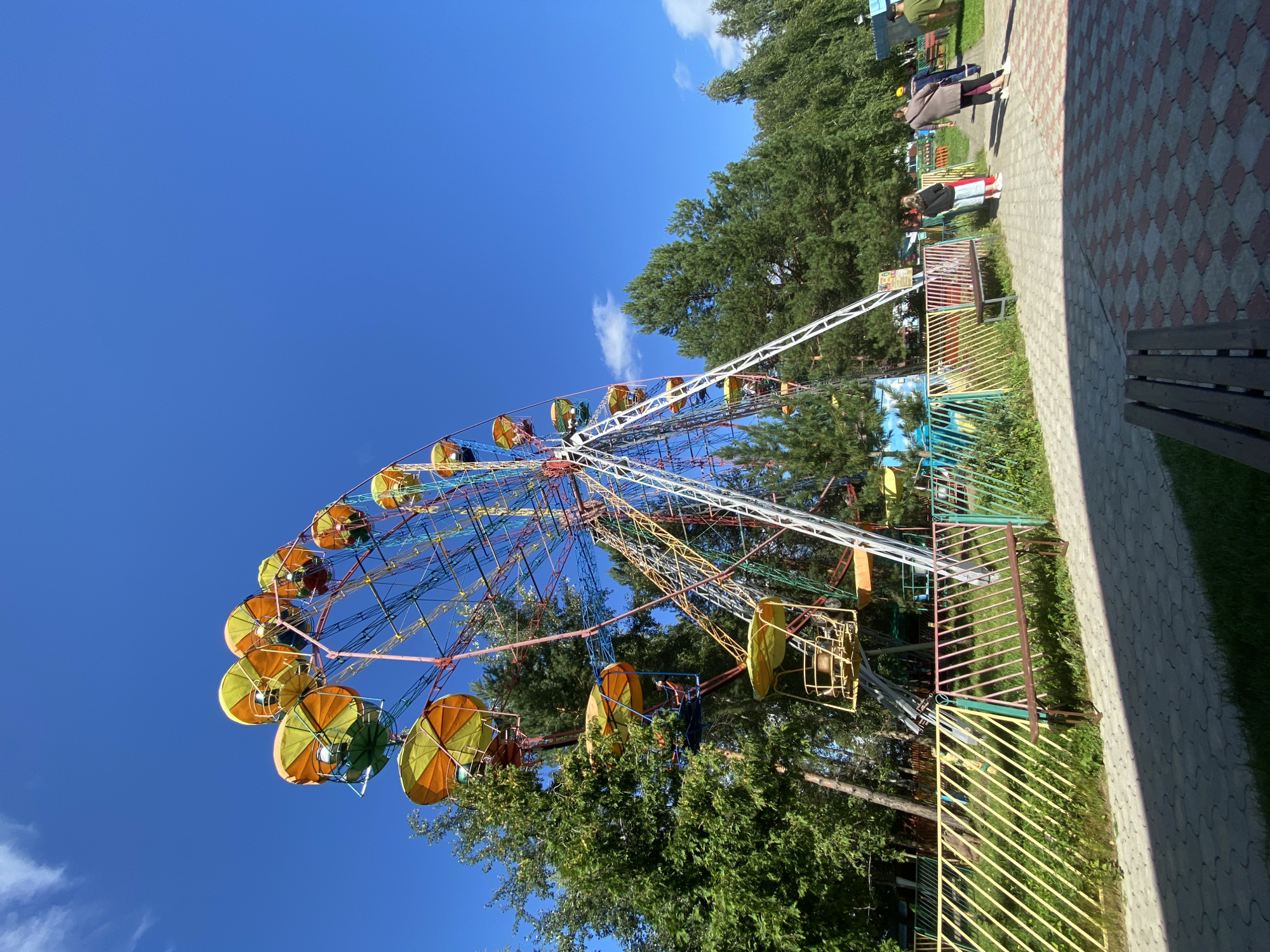 The image depicts a Ferris wheel at an amusement park, likely located in Chaikovsky, Russia, under a bright blue sky with scattered clouds. The Ferris wheel is multi-colored, with yellow and orange canopies on its gondolas, and is surrounded by lush green trees. In the foreground, a paved walkway with a diamond pattern in red and grey is visible, bordering a fence made of colorful metal poles. A wooden bench sits by the fence. Several people are seen walking on the pathway, some appearing to be visitors to the park. The overall scene suggests a sunny day at a recreational area.