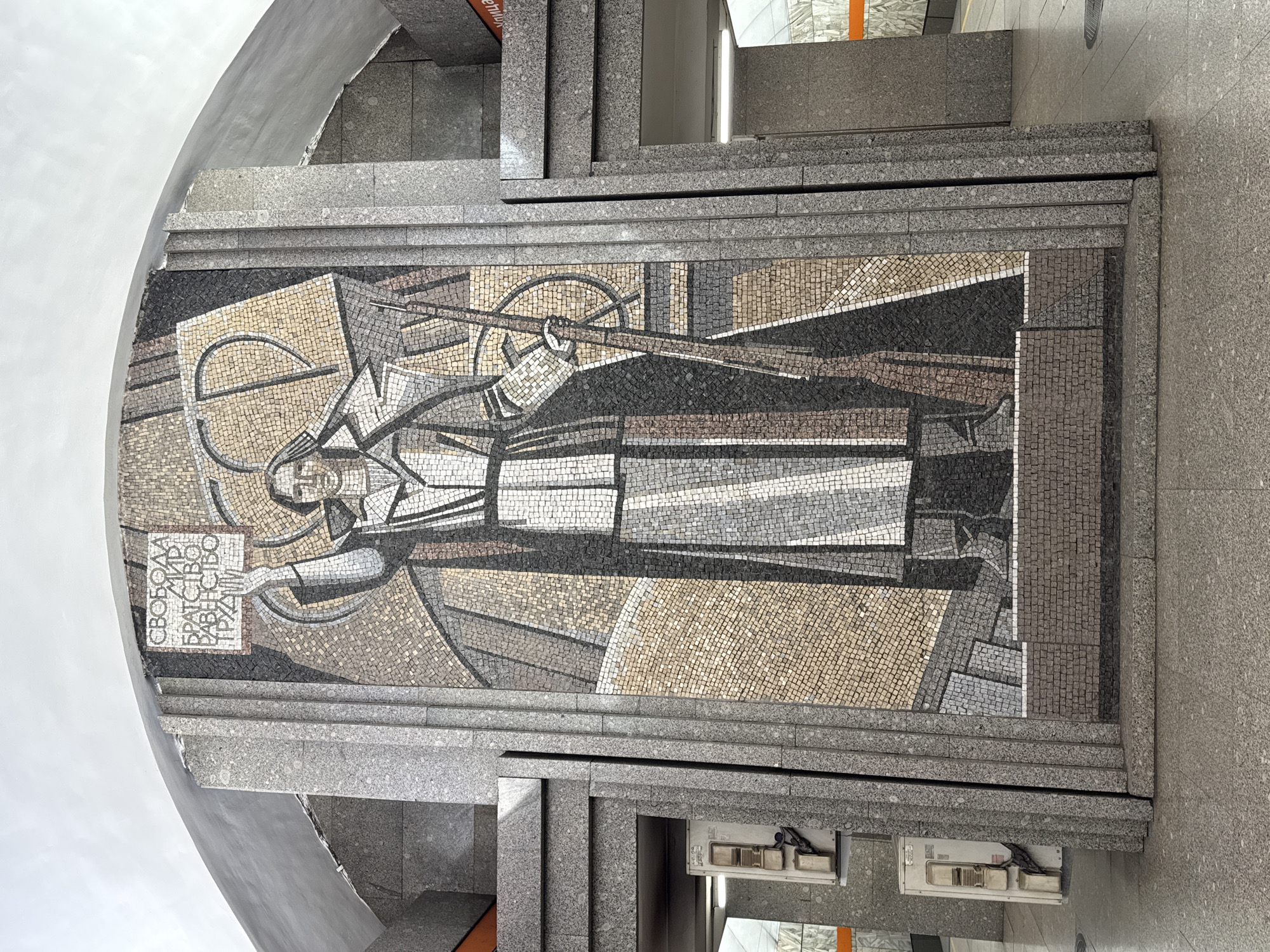 The image shows a large mosaic mural in a public space, likely a metro station, in Saint Petersburg, Russia. The mural is stylized in a socialist realist manner, depicting a worker figure holding a rifle or tool and a sign with Cyrillic text. The mosaic is constructed of small tiles in various shades of gray, beige, white, and black, creating a textured and somewhat abstract image. The mosaic is set within a structure of gray stone, possibly granite, that includes architectural elements such as steps or ledges. The environment suggests an underground location.