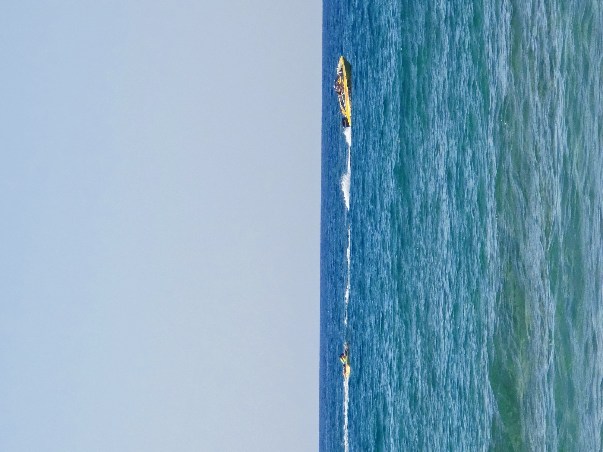 An eye-level, medium shot shows two watercraft on the bright blue ocean under a clear sky. The water is choppy, with white foam trailing behind both vessels. In the distance to the left, a single person is riding a jet ski. Closer to the center and further to the right, a yellow speed boat carries several passengers. The horizon line is sharp, dividing the sea from the sky. The image appears to be taken from a shore in Malia Municipal Unit, Greece.