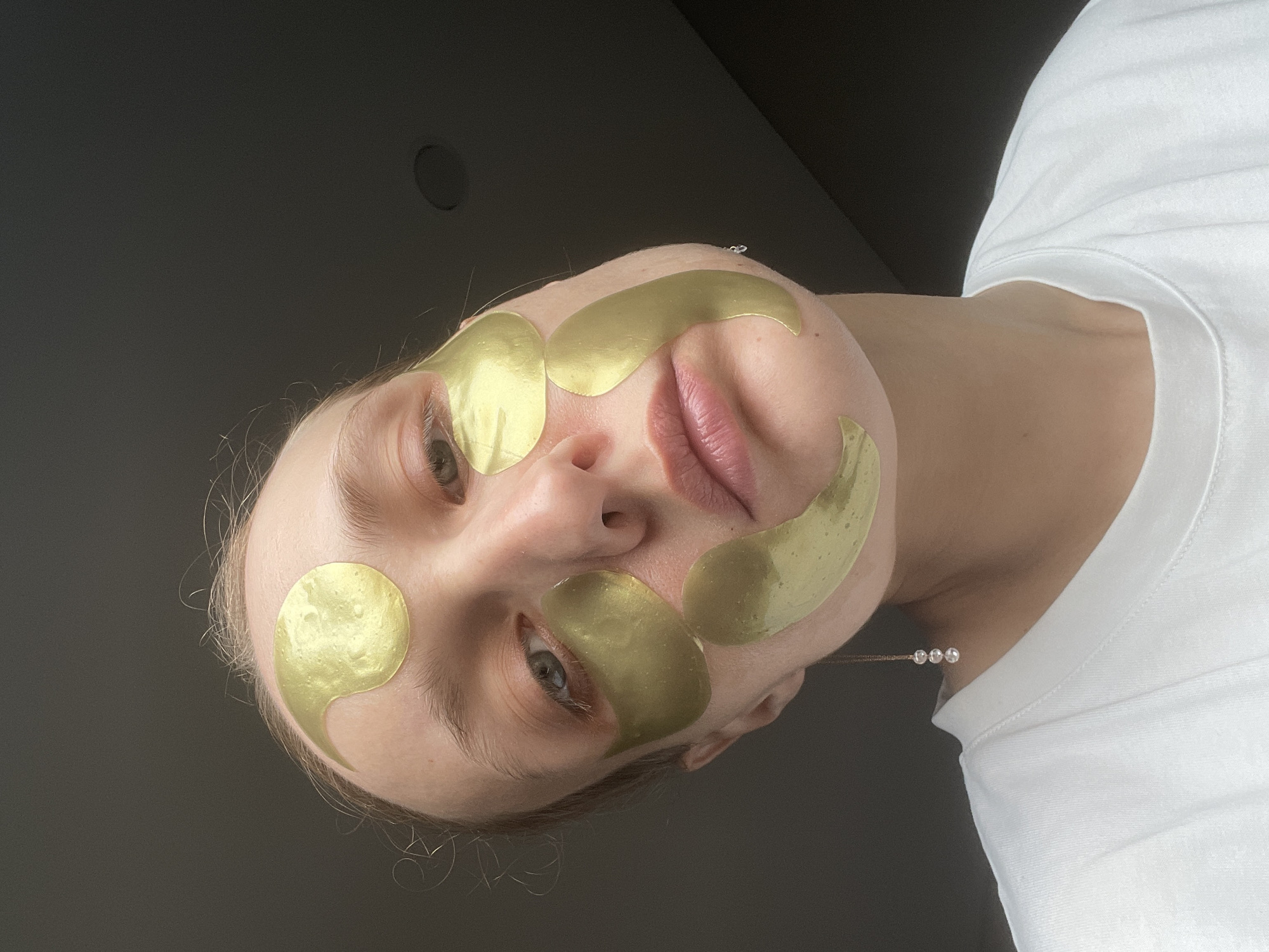 A person with fair skin is pictured from a low angle, with a dark grey ceiling visible in the upper portion of the frame. The person has golden facial patches applied to their forehead, cheeks, and under their eyes. The patches are slightly curved in shape. The person is wearing a white t-shirt. A delicate necklace with small, round beads is visible on the person's neck. There is no indication that the image was taken in the city of Podshivalovo, Russia.