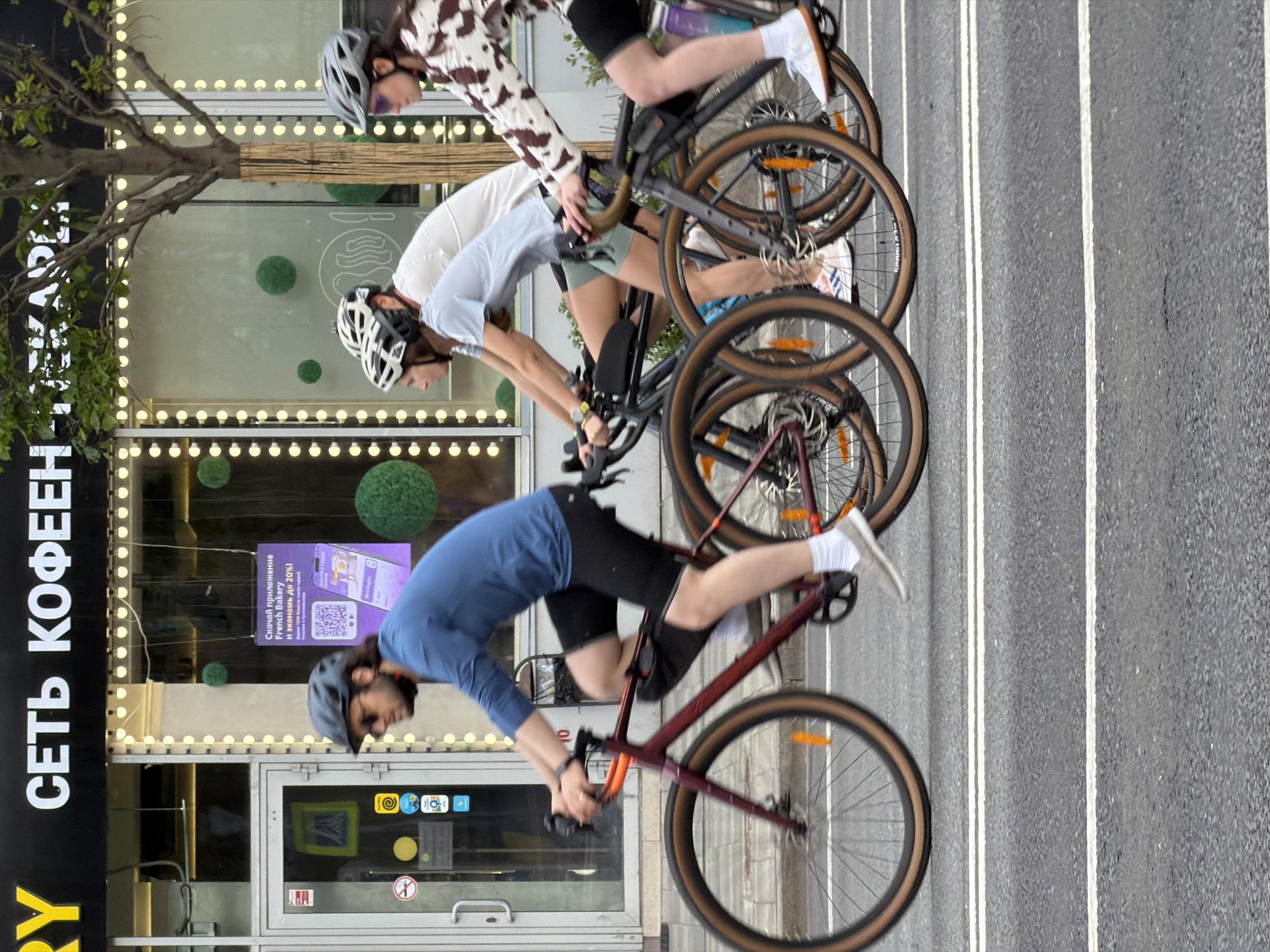 The image shows three people riding bicycles in Moscow, Russia. They are riding in a single file line on a city street with white lane markings. The cyclist in the front is wearing a blue shirt, black shorts, and a dark helmet. The second cyclist is wearing a white shirt, light green shorts, and a white helmet. The third cyclist is wearing a white and brown cow-patterned shirt, black shorts, and a grey helmet. The building behind them is a "Сеть Кофеен" (coffee chain) with a sign in Russian and English promoting a French Bakery app. There are decorative green spheres on the building facade.