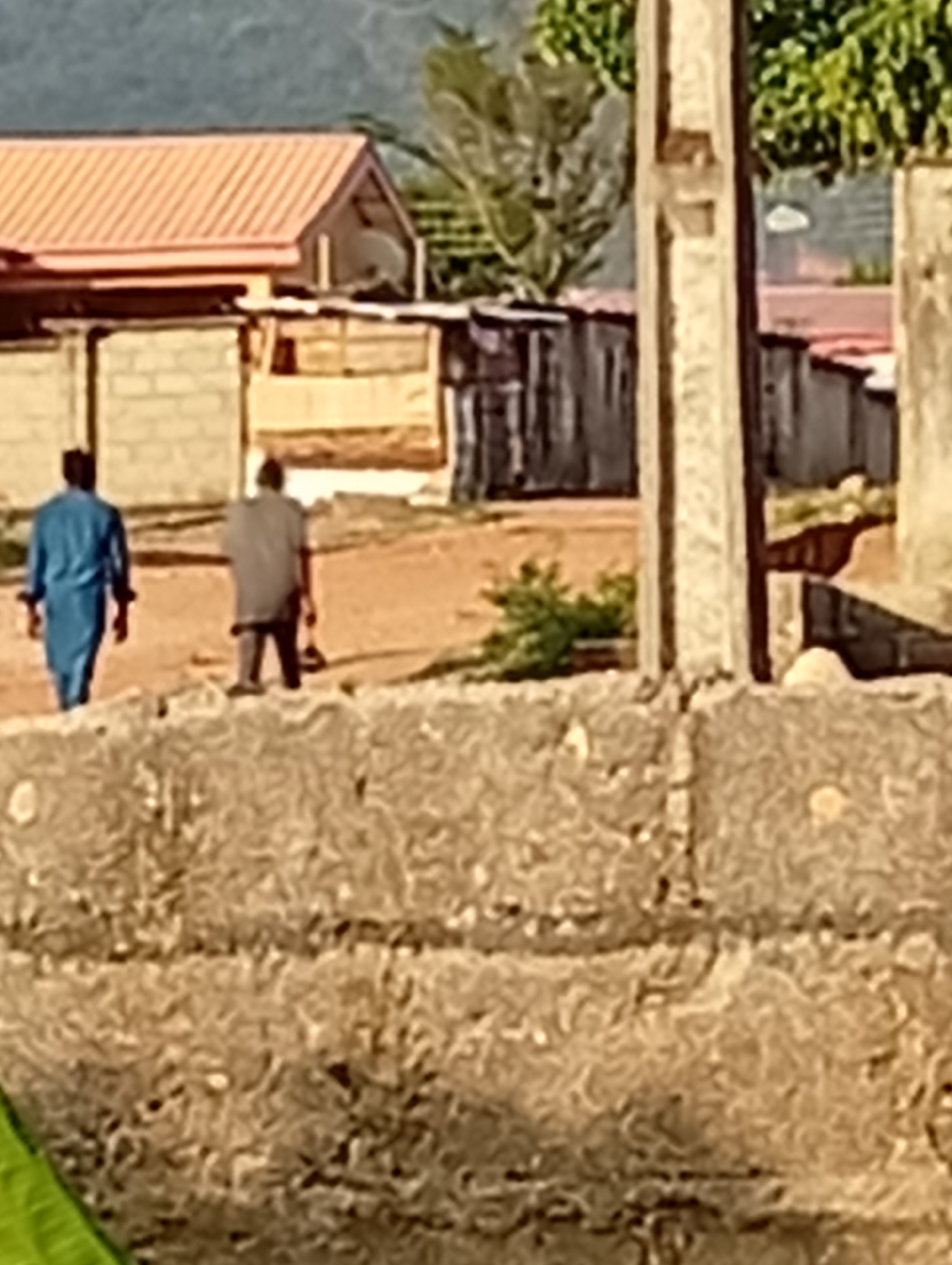 Two people walk down a dirt road in an area with modest dwellings. One person wears a blue outfit, the other a gray shirt. Buildings with red and gray roofs line the road. Vegetation is present in the background. A concrete structure is in the foreground. The location is a settlement within the city of Unknown, Unknown.