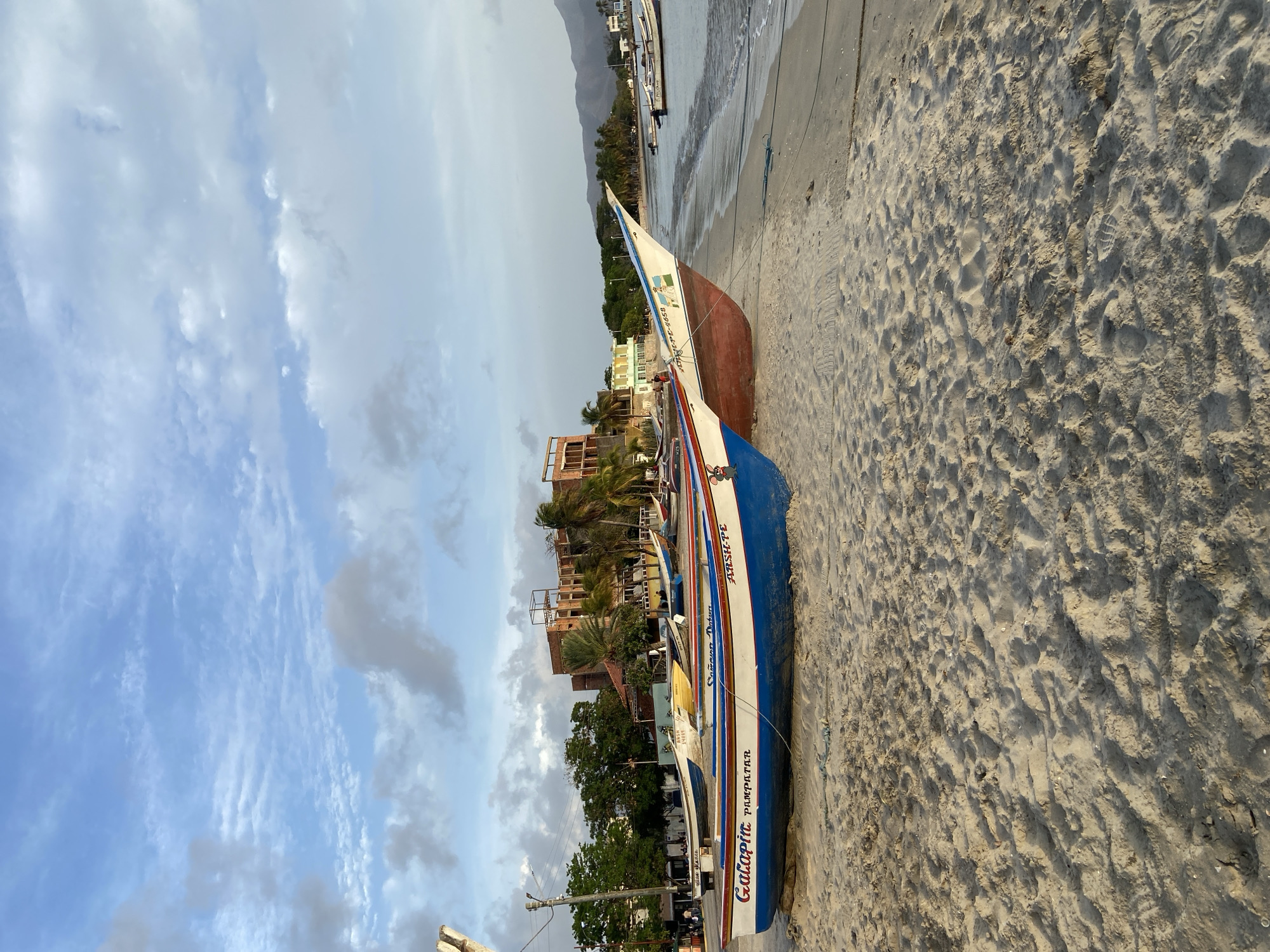 The image depicts a beach scene in Juan Griego, Venezuela, with several colorful fishing boats resting on the sand near the water's edge. The sky is partly cloudy, with a mix of white and gray clouds against a pale blue background. In the background, there are buildings, palm trees, and mountains in the distance. The sand is textured with footprints and some debris. The boats are the primary focus, with some bearing names and markings. One boat in the foreground is blue and white with red trim, labeled "Galapita Pampatar." Another boat is white and red, and a yellow boat is visible behind them. The water is calm, with gentle waves lapping at the shore. Several other boats are anchored further out in the bay. The overall impression is a peaceful, coastal setting.