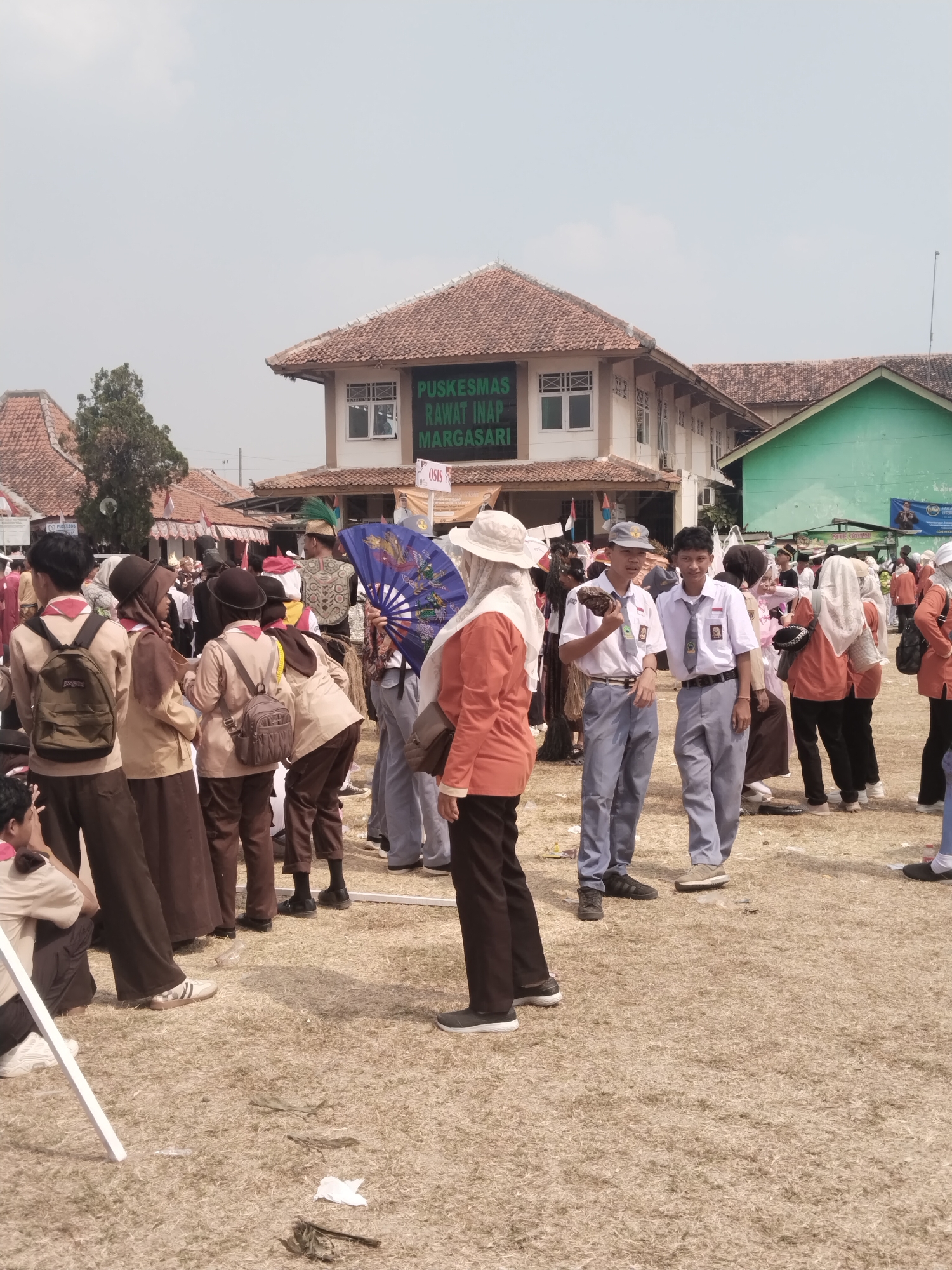 The image depicts an outdoor scene in front of a building identified as "PUSKESMAS RAWAT INAP M<bos>ARGASARI," a health center in Margasari, Indonesia. A gathering of people, many of whom appear to be students in uniform, are present on a dry, grassy field. Some individuals are wearing khaki uniforms with brown pants and some are wearing white shirts with grey ties and pants. Several people are holding or interacting with traditional fans, one of which is a decorative blue fan with intricate designs. A few individuals are also holding what appear to be snacks. The background features tiled-roof buildings, including the prominent Puskesmas building and a bright green structure. The sky is overcast. The overall impression is of a community event or school activity taking place in the afternoon.
