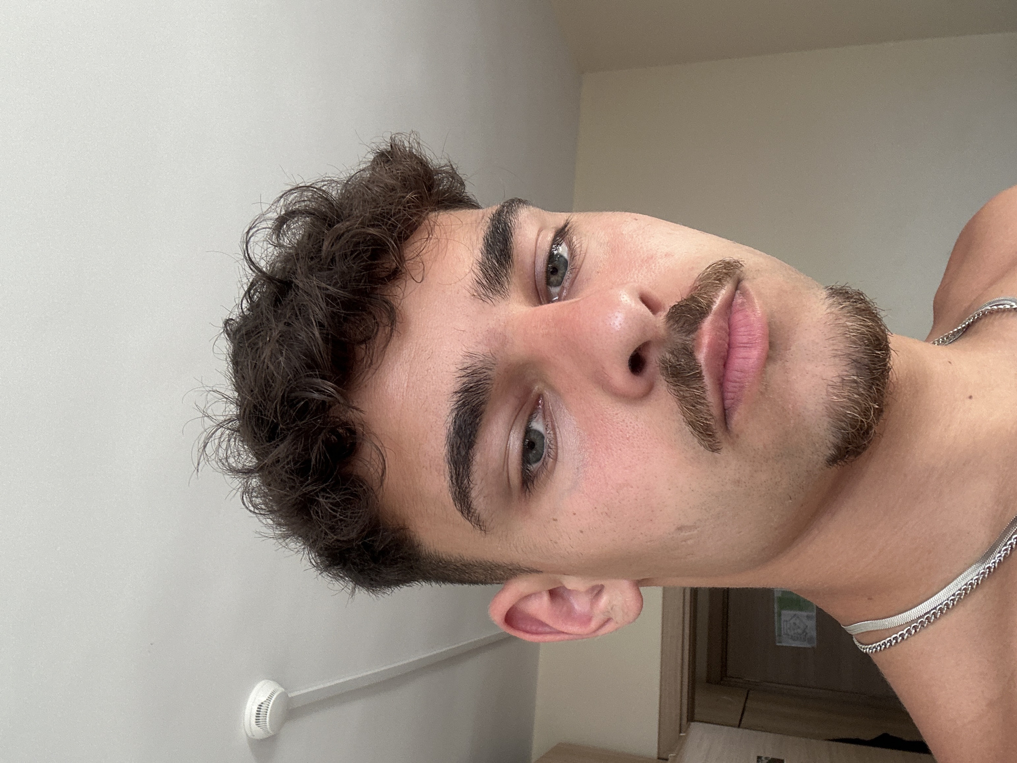 A close-up, tilted headshot of a young male with curly brown hair, light skin, and hazel eyes. He has a mustache and a goatee. He is wearing a silver chain necklace. The background consists of a pale gray wall, a white smoke detector, and a glimpse of a door. The location is not discernible based on the image content. Sofia, Bulgaria cannot be determined from the information in the image.