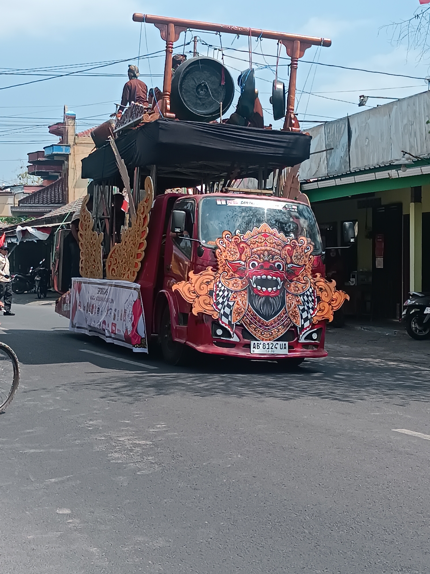The image depicts a red truck adorned with a large, colorful Balinese Barong mask on its front. The truck is participating in a procession or parade, as indicated by the banner draped on its side and a wooden structure with gongs mounted on its roof, suggesting a musical performance. Several individuals are visible: one person is seated on the roof structure, seemingly involved with the musical instruments, while another person stands near the side of the truck, dressed in a tan uniform, possibly a security or event staff member. In the background, buildings with typical Indonesian architecture, including tiled roofs and awnings, line a street. The sky is clear and blue, suggesting daylight. The location is identified as Imogiri, Indonesia, a known cultural and historical area. The scene appears to be taking place on a paved road in what seems to be a commercial or residential district. The overall atmosphere suggests a lively cultural event or celebration.