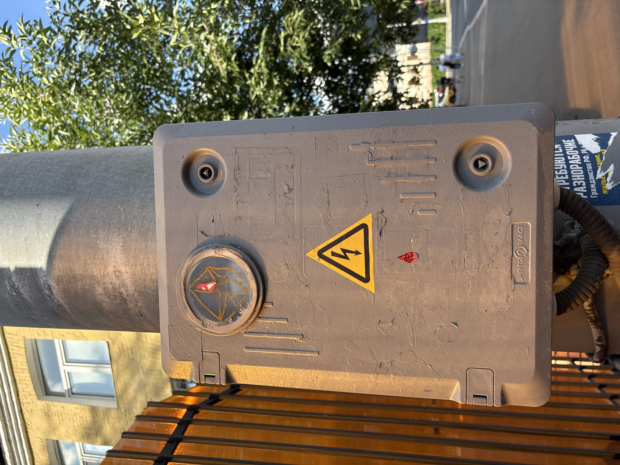 An outdoor, close-up, eye-level shot focuses on a gray utility box mounted on a concrete pole in Moscow, Russia. The box is weathered and has several marks and scratches. Prominently displayed on the box is a yellow triangular warning sign with a black lightning bolt symbol. Above this sign, a circular window shows an abstract red and yellow graphic. Below the warning sign, a small red, tear-drop shaped sticker is visible. Text, partially obscured, is present on the box, including what appears to be a logo reading "ЭНЕРГО ПЛАСТ" and additional Cyrillic text at the bottom. The pole also has a corrugated conduit entering the utility box from below. To the left of the pole, a portion of an orange, translucent fence runs vertically. In the background, a yellow building with white-framed windows is partially visible, along with green trees and a bright blue sky. Further in the background, a street scene shows a sidewalk, a road, and several blurred figures walking. The overall lighting suggests it is daytime, possibly late afternoon, as indicated by the warm light casting shadows.