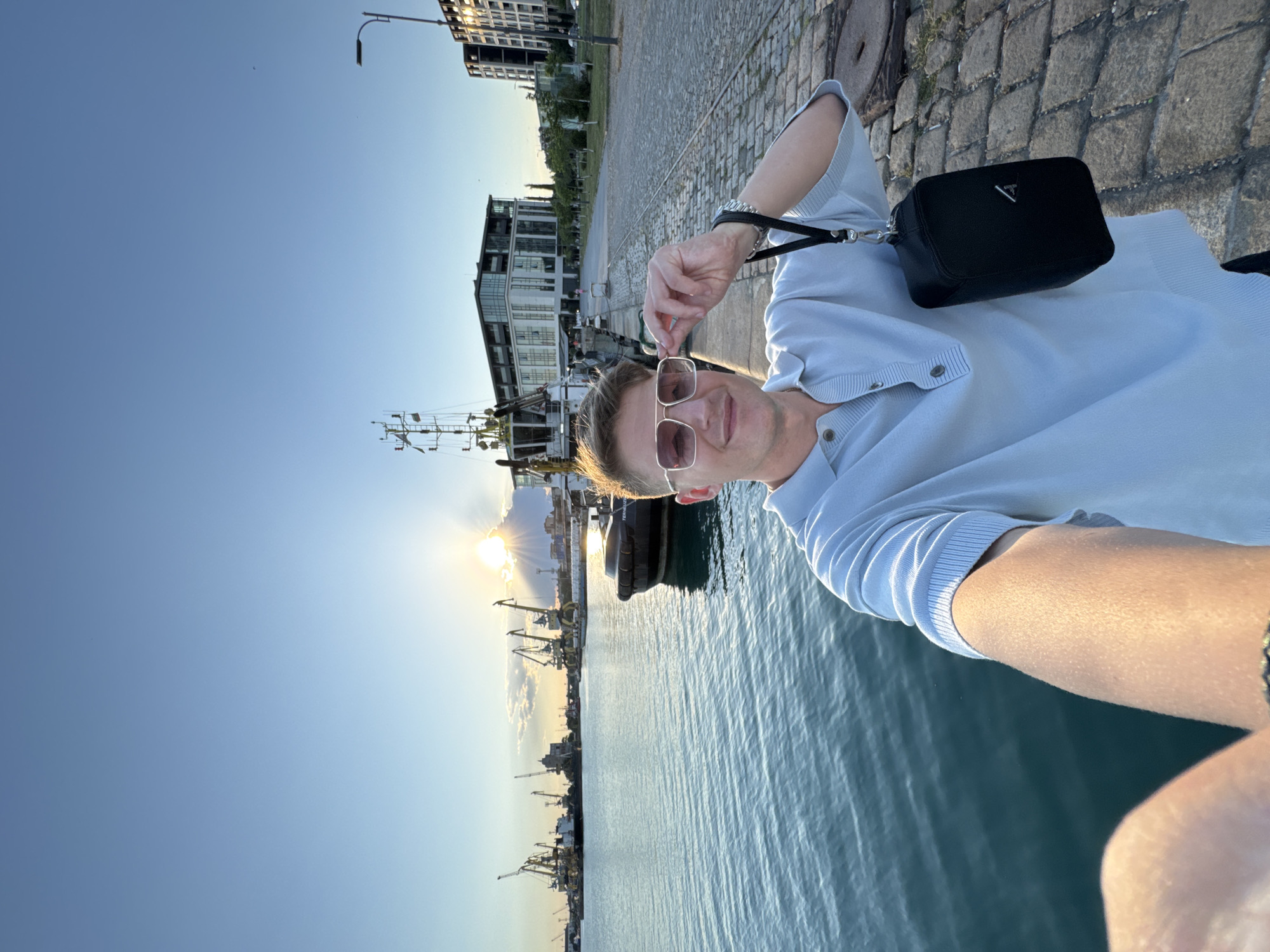 A young man, wearing sunglasses and a light blue polo shirt, takes a selfie in the port of Burgas, Bulgaria, with the sea and industrial cranes in the background. The sun is setting, casting a warm glow over the scene. The man is holding his sunglasses with his right hand, and a black pouch is hanging from his wrist. The port appears active, with several boats and ships docked. The city skyline is visible in the distance, with modern buildings. The ground is paved with cobblestones.