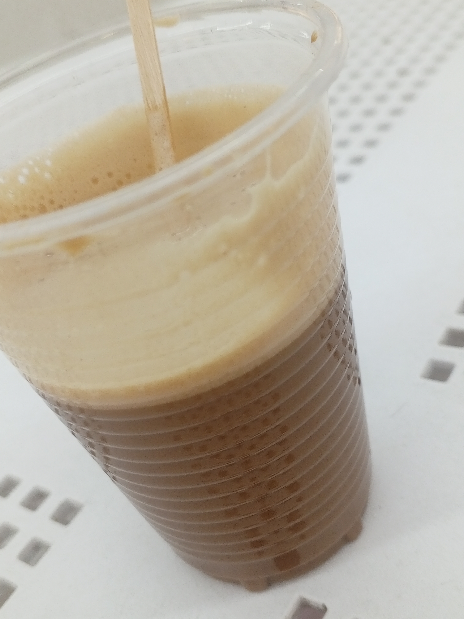 A close-up, angled shot features a beverage in a clear plastic cup, likely taken indoors. The drink consists of two distinct layers: a dark brown liquid occupying the lower portion of the cup, and a thick, light tan, frothy foam on top with small bubbles. A thin, light-colored wooden stirring stick is positioned vertically within the drink, submerged in the foam, suggesting it is either being mixed or is ready for consumption.
The plastic cup is ribbed with horizontal ridges on its exterior. It is placed on a white or very light gray surface that displays a repeating grid pattern of small, recessed squares. This textured surface extends across the background, with shadows indicating bright, possibly overhead lighting. No people are visible in the image, and there is no discernible text. The scene is set in Porlamar, Venezuela.