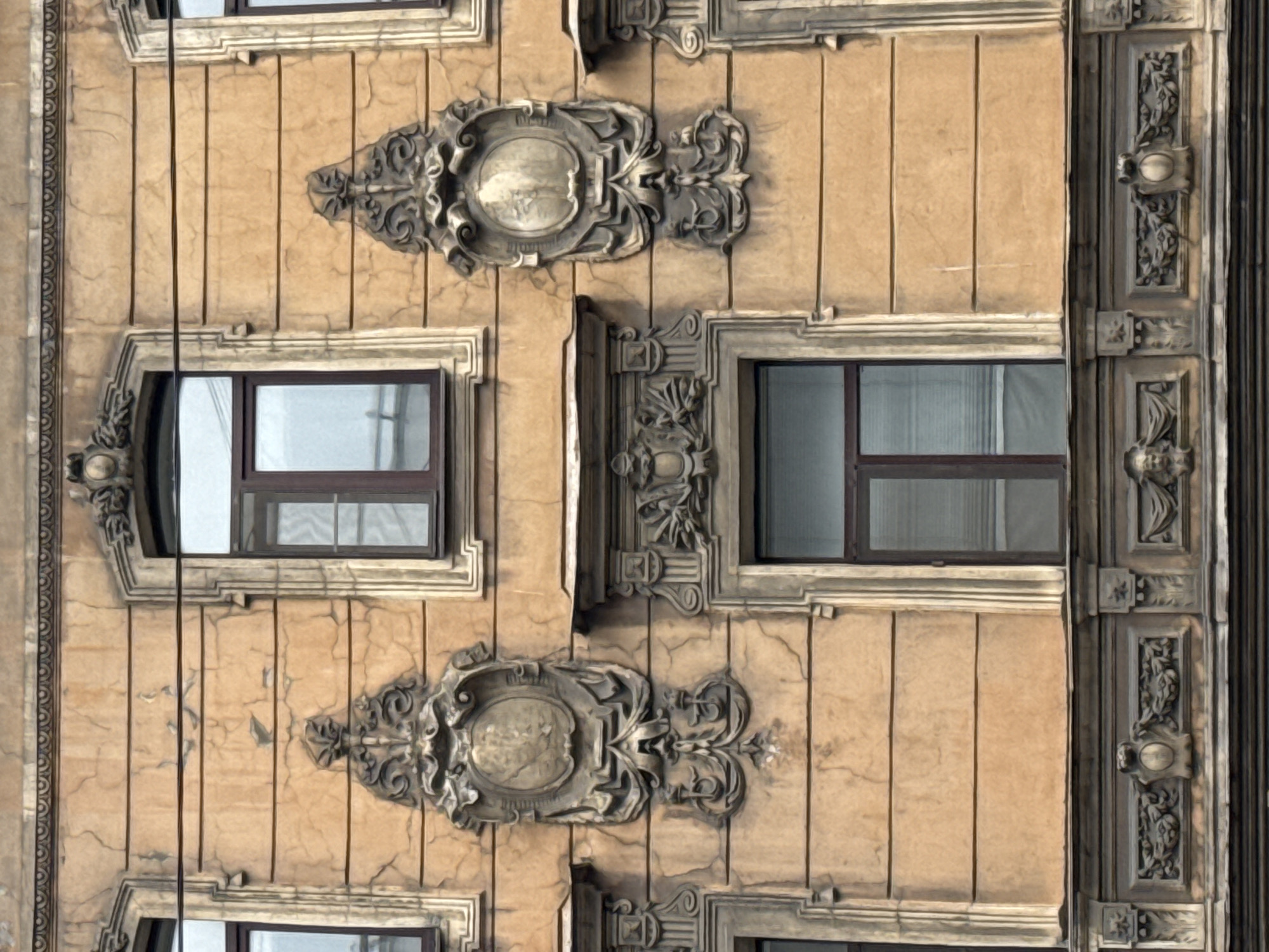 The image shows a section of a building facade in Saint Petersburg, Russia. The architecture features detailed ornamentation around the windows and decorative panels between them. The facade has a tan-colored textured finish, and the windows are dark-framed with reflective glass. The design includes classical-style embellishments and vertical pilasters. The overall style appears to be representative of historic architecture, possibly from the late 19th or early 20th century.