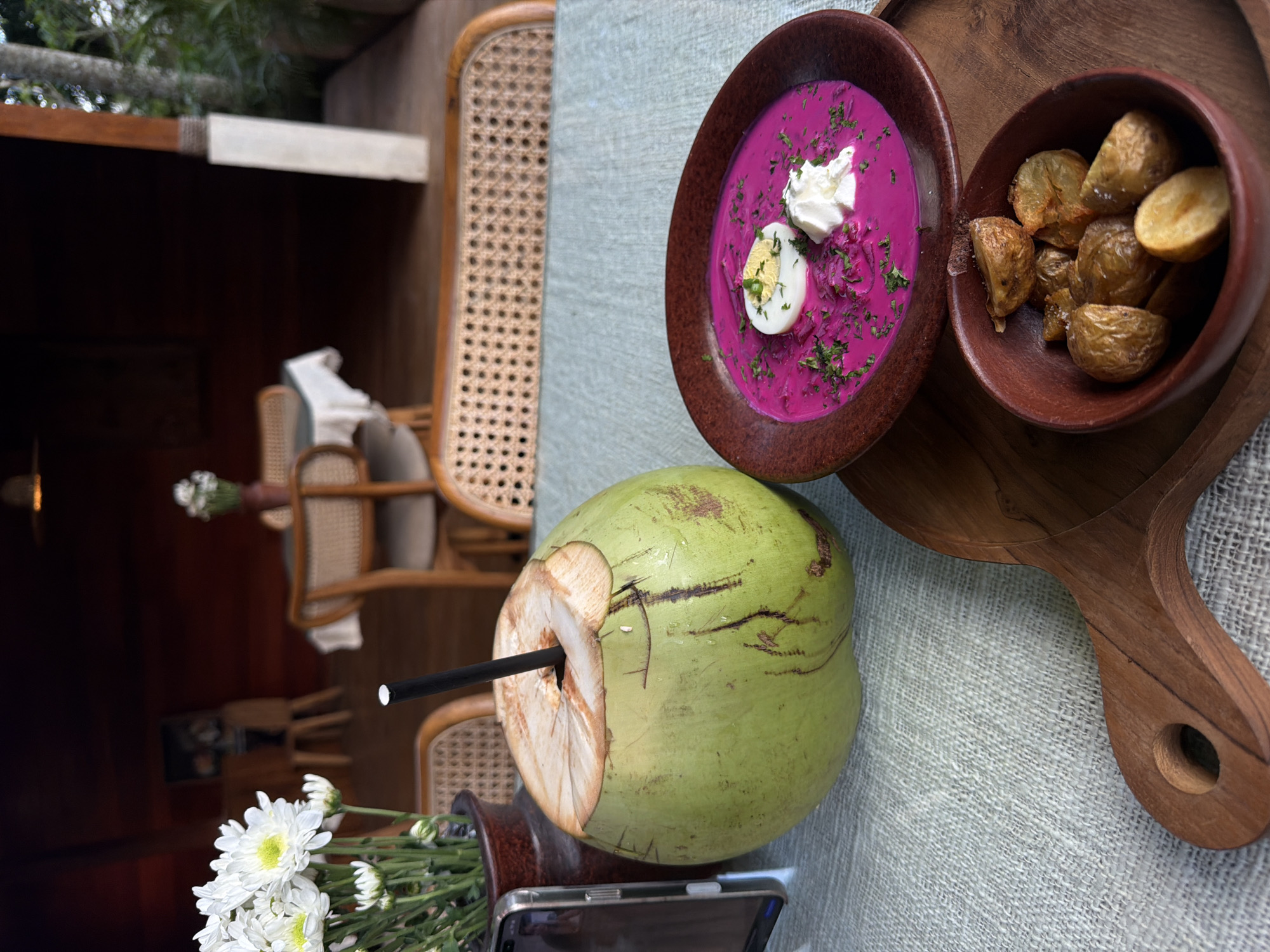 The scene presents a dining table setting in Petulu, Indonesia, focusing on a culinary presentation. On a light-colored, textured tablecloth, a wooden serving board holds two earthenware bowls. One bowl contains a bright pink soup, possibly beetroot-based, garnished with a dollop of white cream, halved hard-boiled egg, and chopped herbs. The second bowl contains small, roasted potatoes.
Next to the board, a green coconut with a straw inserted sits atop a dark vase containing white daisies. A smartphone is positioned beneath the coconut.
A woven rattan chair is visible in the foreground and background, suggesting an outdoor or semi-outdoor dining setting. Other tables and chairs are present in the background. The lighting appears natural, and the background is partially obscured by greenery.