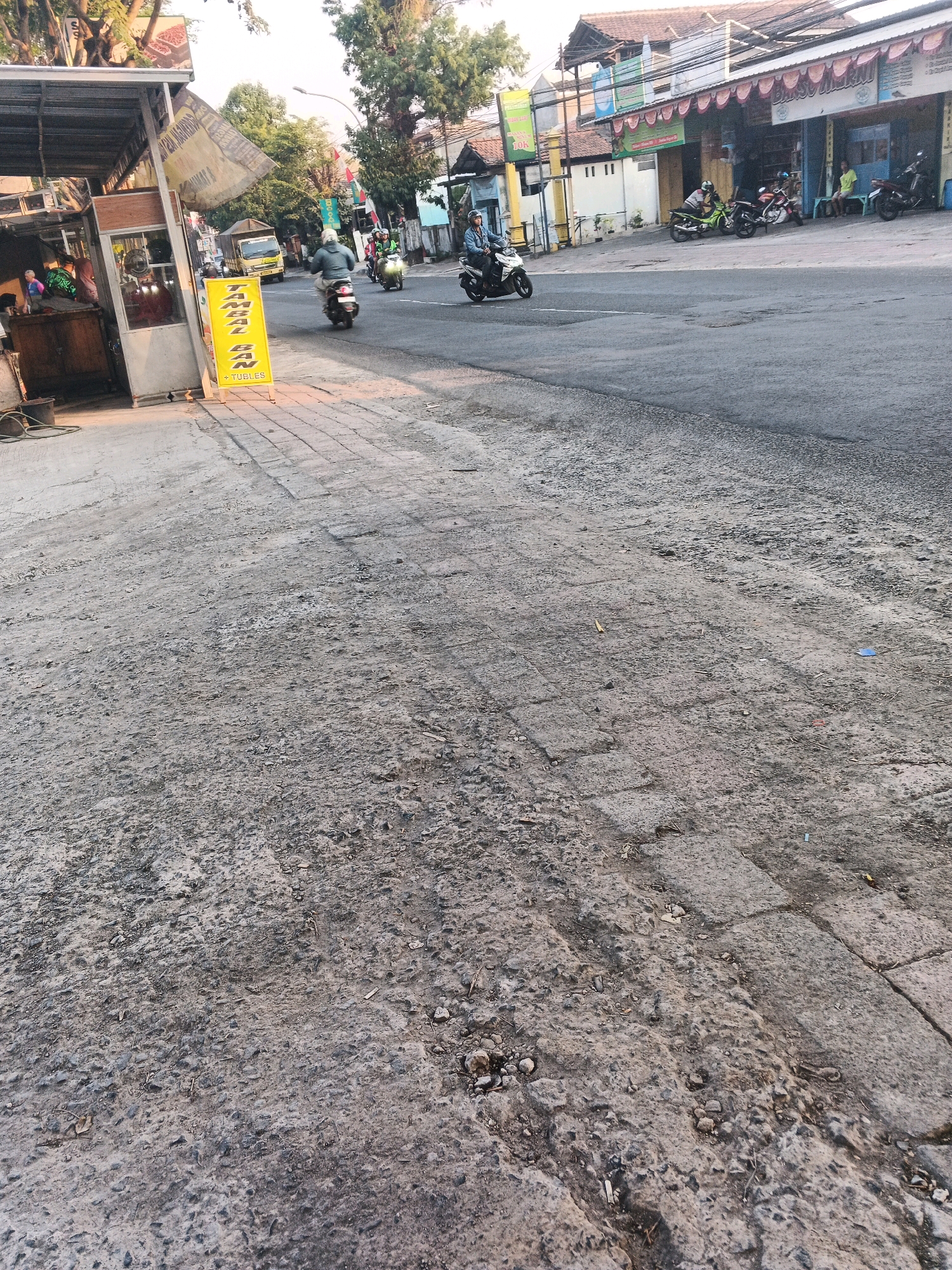 The image depicts a street scene in Ungaran, Indonesia, likely during the day given the lighting. In the foreground, a cracked and weathered concrete sidewalk runs along the left side of the frame, with some gravel and debris scattered on its surface. To the left, a small open-air structure, possibly a food stall, is visible with a metal roof. A yellow sign with red lettering stands prominently on the sidewalk, indicating "Tambal Ban + Tubles" (Tire Patching + Tubeless), suggesting a tire repair service. Several people are visible within and around the stall.
The street itself is paved with asphalt and has visible lane markings. Traffic moves along the street, with several motorcycles and a truck visible. On the right side of the street, buildings line the road, some with awnings and signs. Indonesian flags are displayed on poles in front of some establishments. The environment suggests an urban or semi-urban setting with typical street infrastructure and commercial activity. The overall impression is one of a busy, everyday street scene.