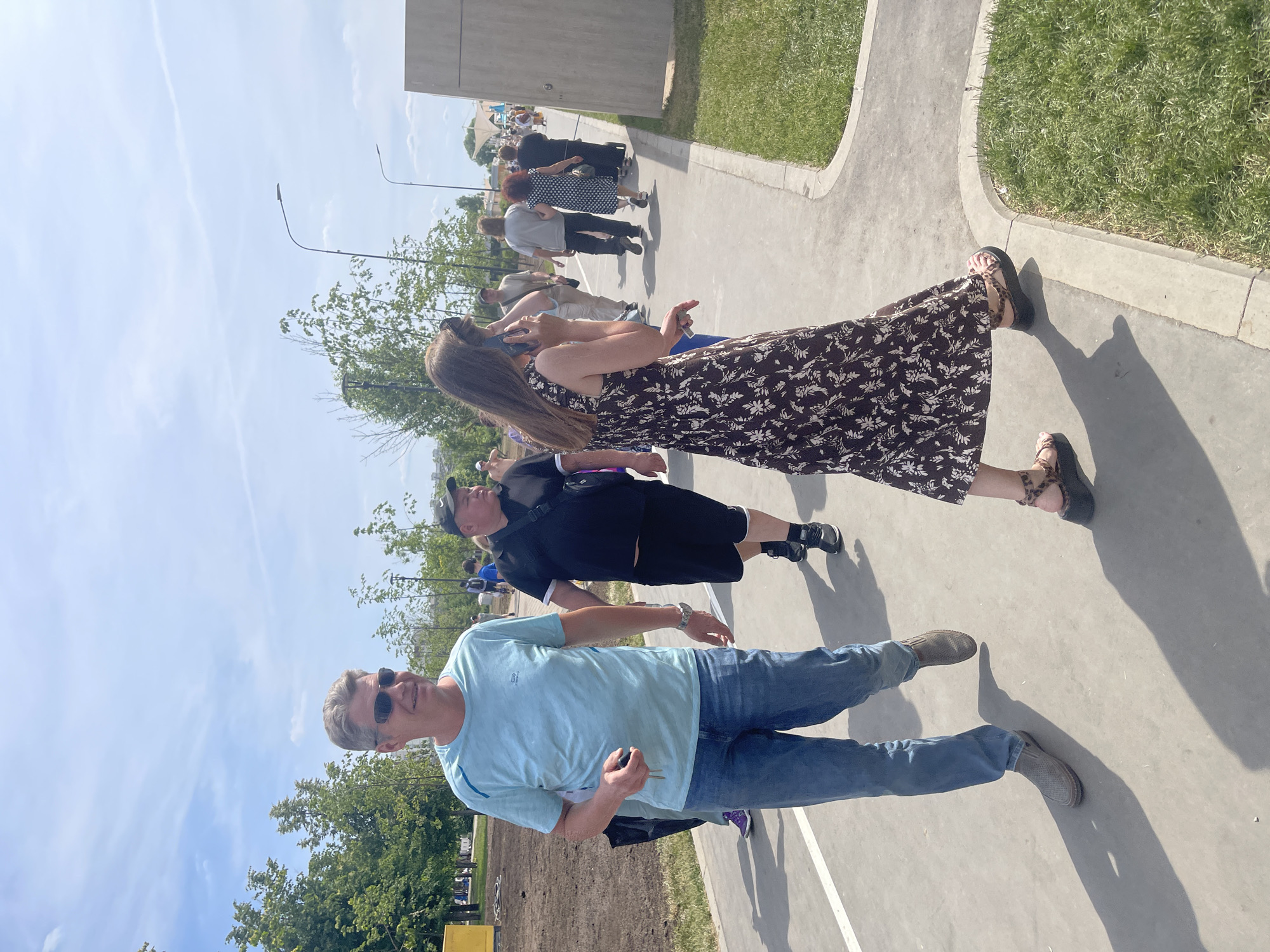 The image depicts an outdoor scene on a sunny day in a modern public park in Moscow, Russia. A grey paved pathway, marked with a white line on its left edge, extends from the foreground into the background, where multiple individuals are walking.
In the immediate foreground, a middle-aged male with short, dark-blonde hair and dark sunglasses walks towards the viewer's left. He wears a light blue short-sleeved t-shirt and blue jeans, with a silver watch on his left wrist. His right hand holds a small, dark object, possibly a car remote or keys. He has a slight smile and looks forward. Adjacent to him on the right, a female with long, dark brown hair walks towards the viewer's right. She is dressed in a dark brown long dress featuring a white floral pattern and wears platform sandals with leopard print straps. Her left hand holds a black smartphone to her ear, indicating she is engaged in a call or interacting with the device.
Behind the foreground figures, several other people are visible walking along the pathway. On the mid-left, a heavy-set male in a black baseball cap, black t-shirt, and black shorts carries a black messenger bag across his chest. Further back on the right, a man in a light grey t-shirt and dark pants, and a woman in a dark polka-dot dress, are observed from behind.
The surrounding environment includes areas of manicured green grass bordering the pathway. Numerous deciduous trees of varying maturity are present; dense foliage is visible on the left, while younger trees are scattered on the right. Several modern streetlights with slender poles and single light fixtures are positioned along the pathway. On the far left, beyond the pathway, an area of disturbed earth and sparse vegetation suggests ongoing landscaping or construction. A large, grey, rectilinear concrete building stands on the far right mid-ground. In the distant background, under a clear blue sky with minimal cloud cover, faint white structures resembling tents or canopies are visible. Strong shadows cast by the people and trees indicate direct sunlight from a high sun angle.