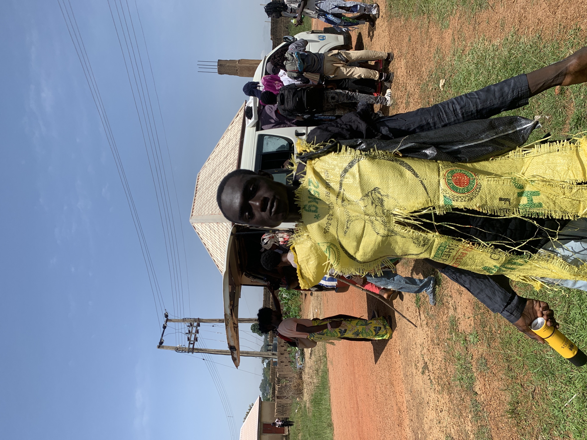 In Gwafan, Nigeria, a man stands in the foreground, wearing a yellow burlap vest over a black shirt and pants. The vest is frayed and appears to be made from a repurposed sack. He holds a yellow cylindrical object, possibly a can or a container, in his left hand. Behind him, a white van with its back door open is parked on a dirt road. A group of people are gathered around the van, some appearing to load or unload items. In the background, there are several buildings, including a structure with a light-colored, tiled roof, and utility poles with electrical wires. The ground is a mixture of dirt and dry grass, indicating an outdoor, possibly rural, setting. The sky is clear and blue.