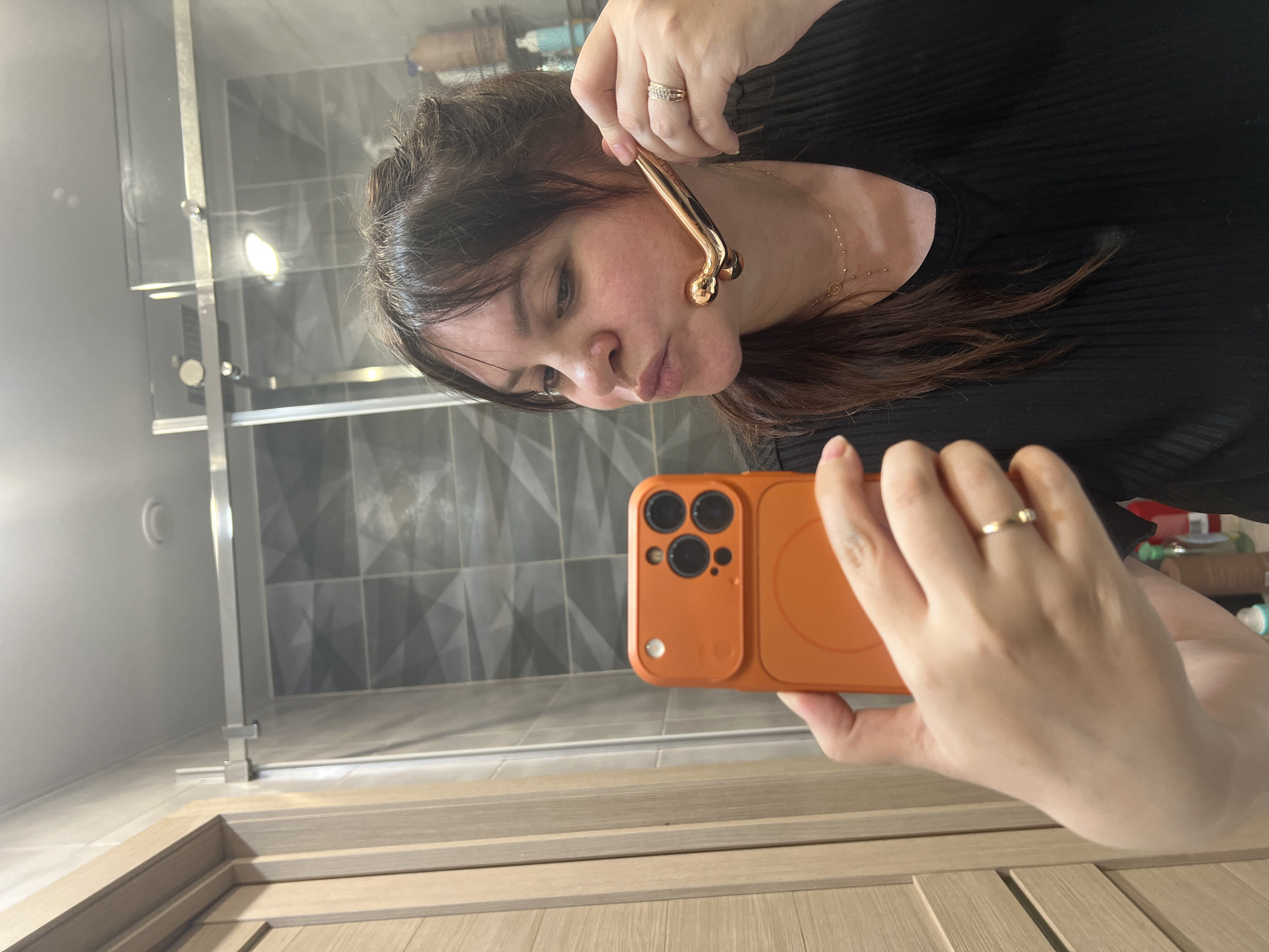 A woman is using a gold facial roller on her face while taking a selfie with an orange cell phone in a bathroom. The bathroom has a shower with gray geometric patterned tiles. The cabinets are made of wood. The location in the city of Noviy, Russia, cannot be determined from the provided image.